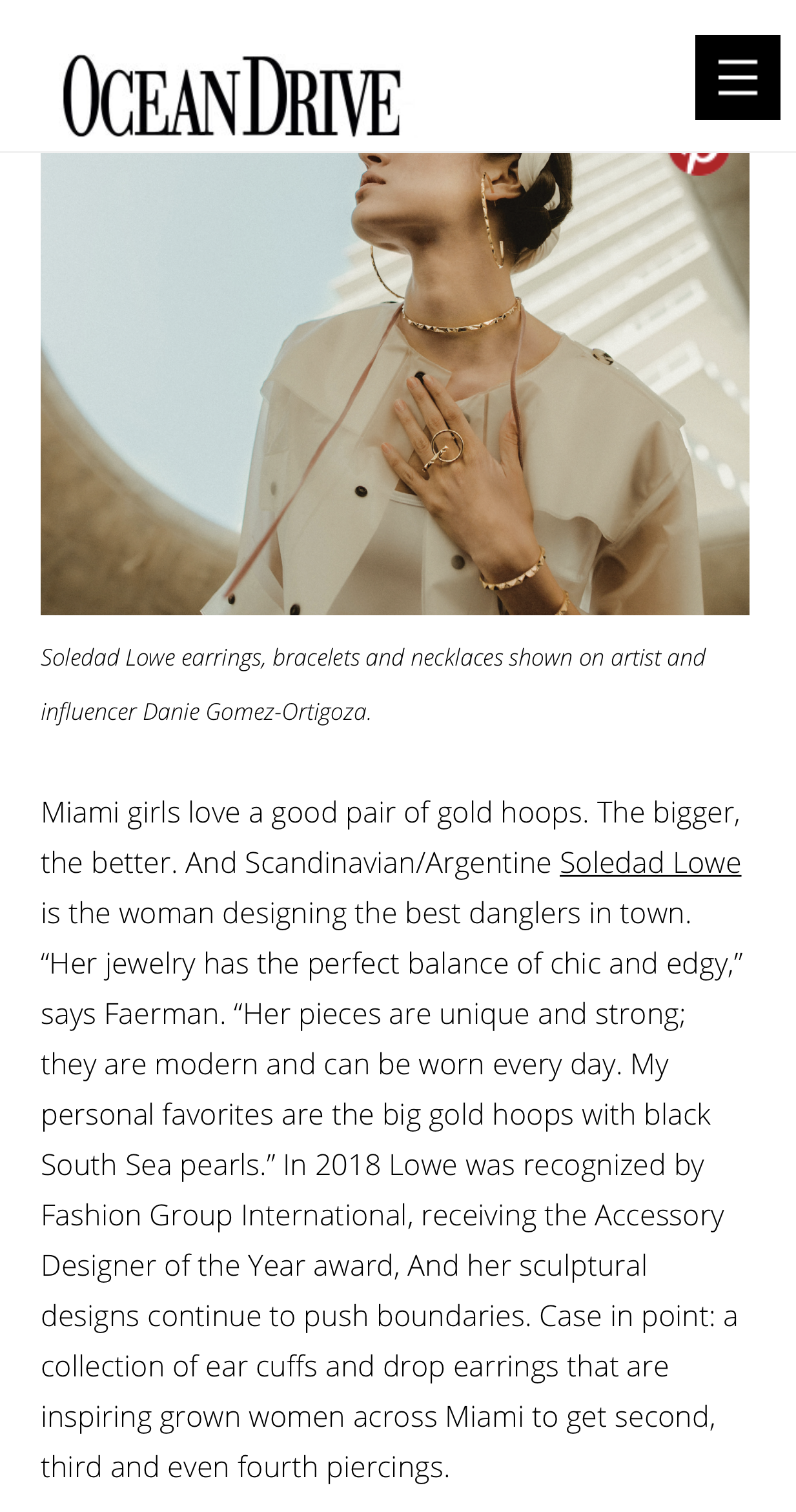 A woman wearing gold jewelry including earrings, bracelets, and necklaces, with her hand placed on her chest. The photo is taken from a low angle, with modern architecture in the background.