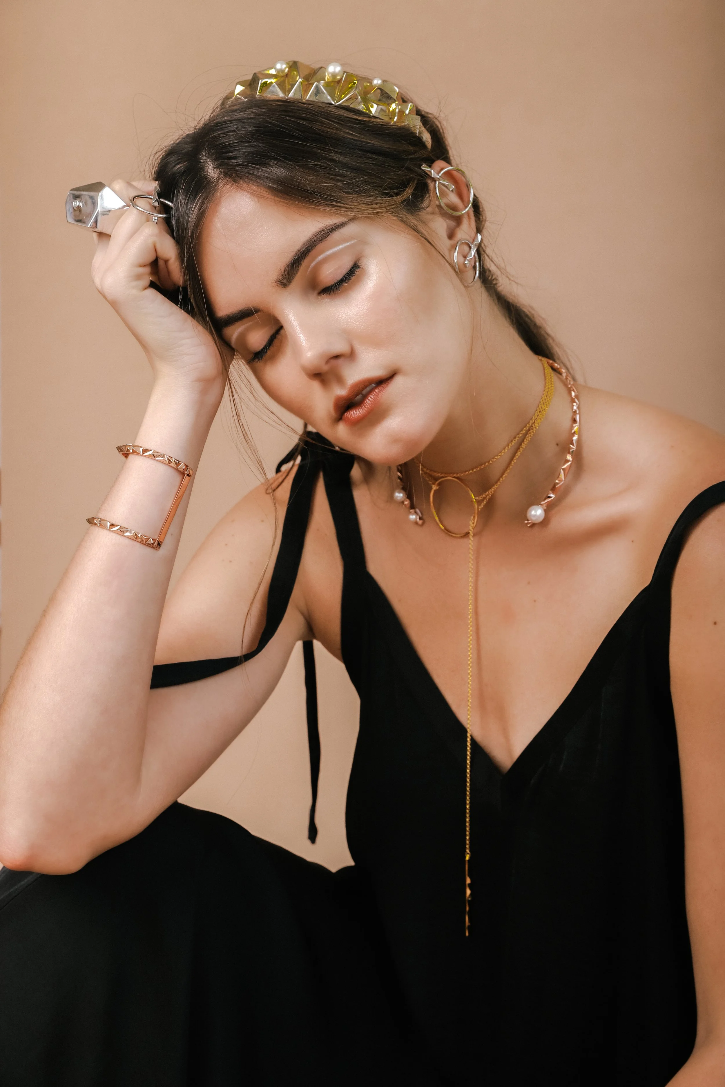 A woman with multiple jewelry pieces, including earrings, necklaces, and bracelets, sits with her eyes closed, resting her head on her hand, wearing a black sleeveless top and gold headband with pearl accents.