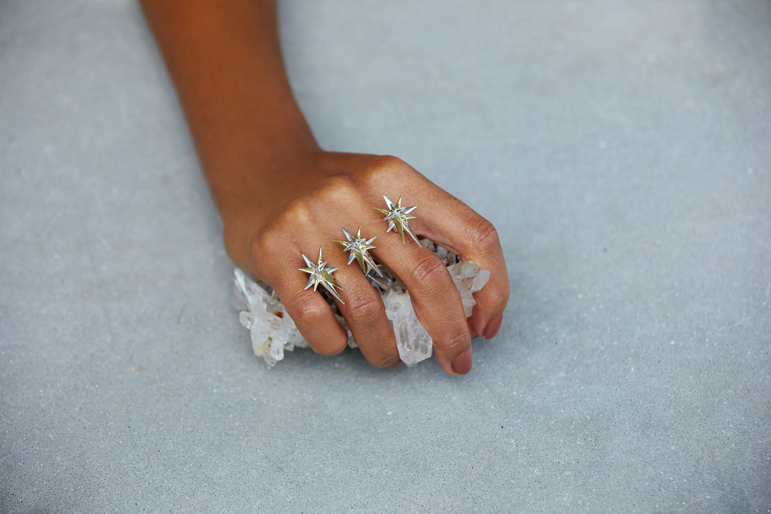A person with dark skin wearing four star-shaped silver rings on their fingers, holding a cluster of clear, crystalline stones, against a light gray background.