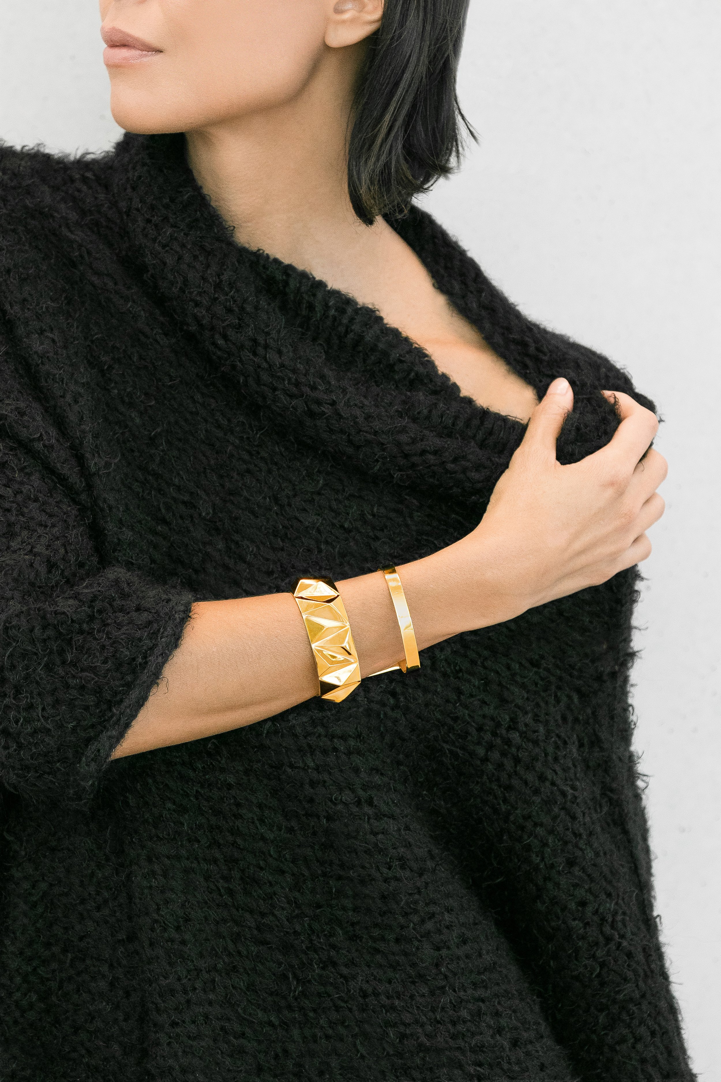 Woman wearing a black textured top and gold jewelry, including two bracelets, with her hand resting on her chest.