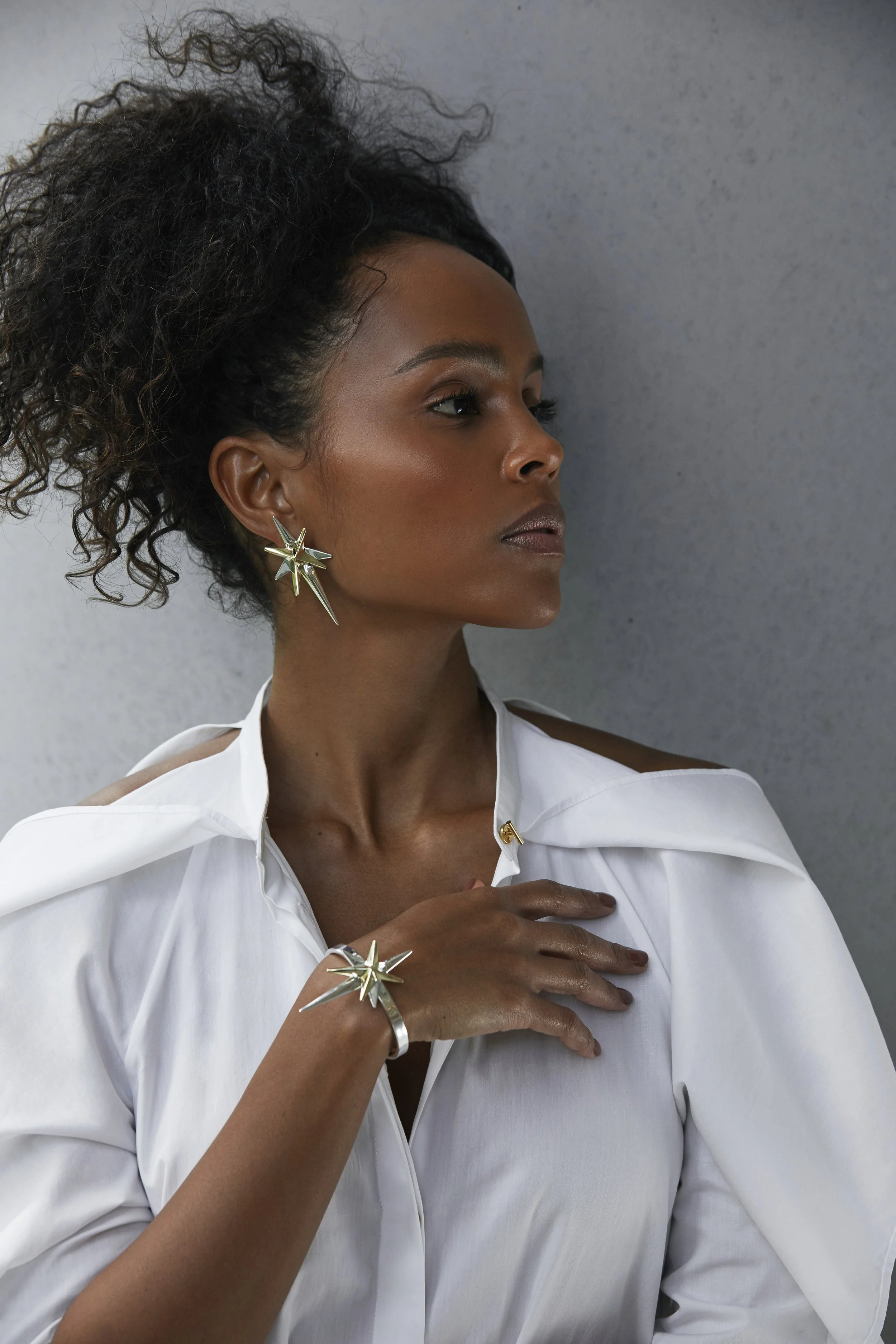 A woman with curly hair wearing statement star-shaped jewelry, a white shirt, and gold accessories, posing against a gray background.