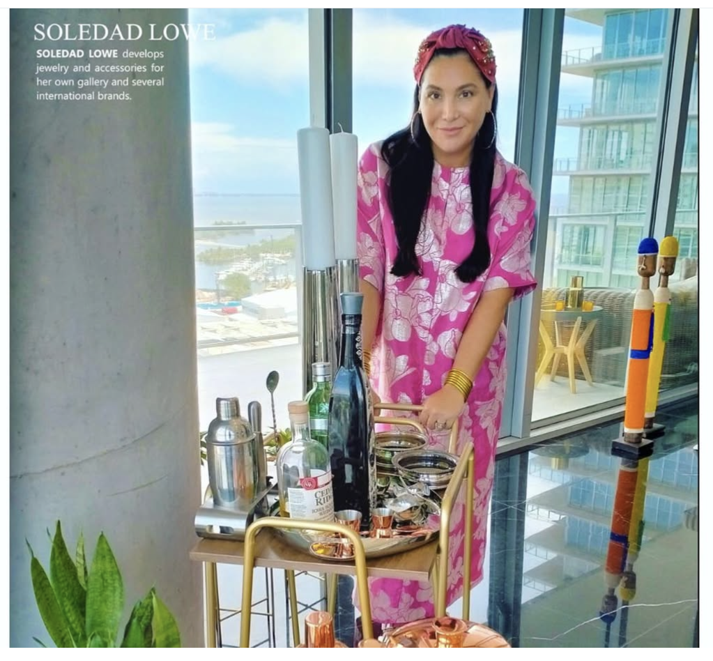 Woman in pink floral dress and headband standing next to a bar cart with bottles and glassware, in a high-rise room with large windows and a city view.