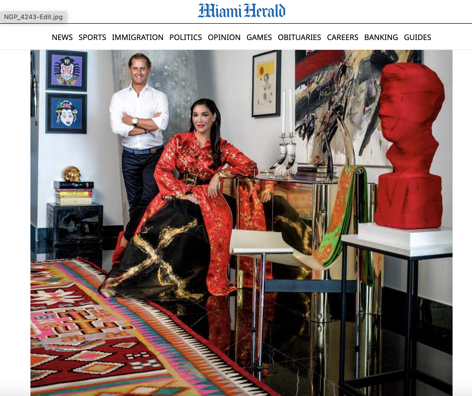 A man and a woman in an art-filled room. The woman is seated, wearing a red and black dress with gold accents. The man stands behind her, wearing a white shirt and dark pants. The room has colorful artwork on the walls, a globe, books, and sculptures