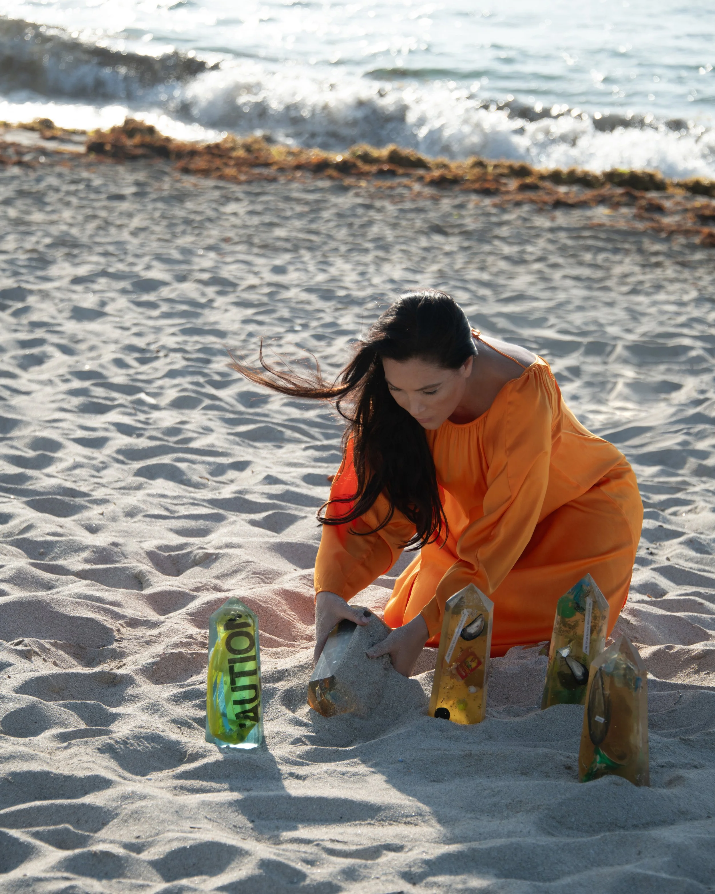 A woman in an orange dress placing sand in plastic bottles labeled with caution symbols on a sandy beach with the ocean waves in the background.