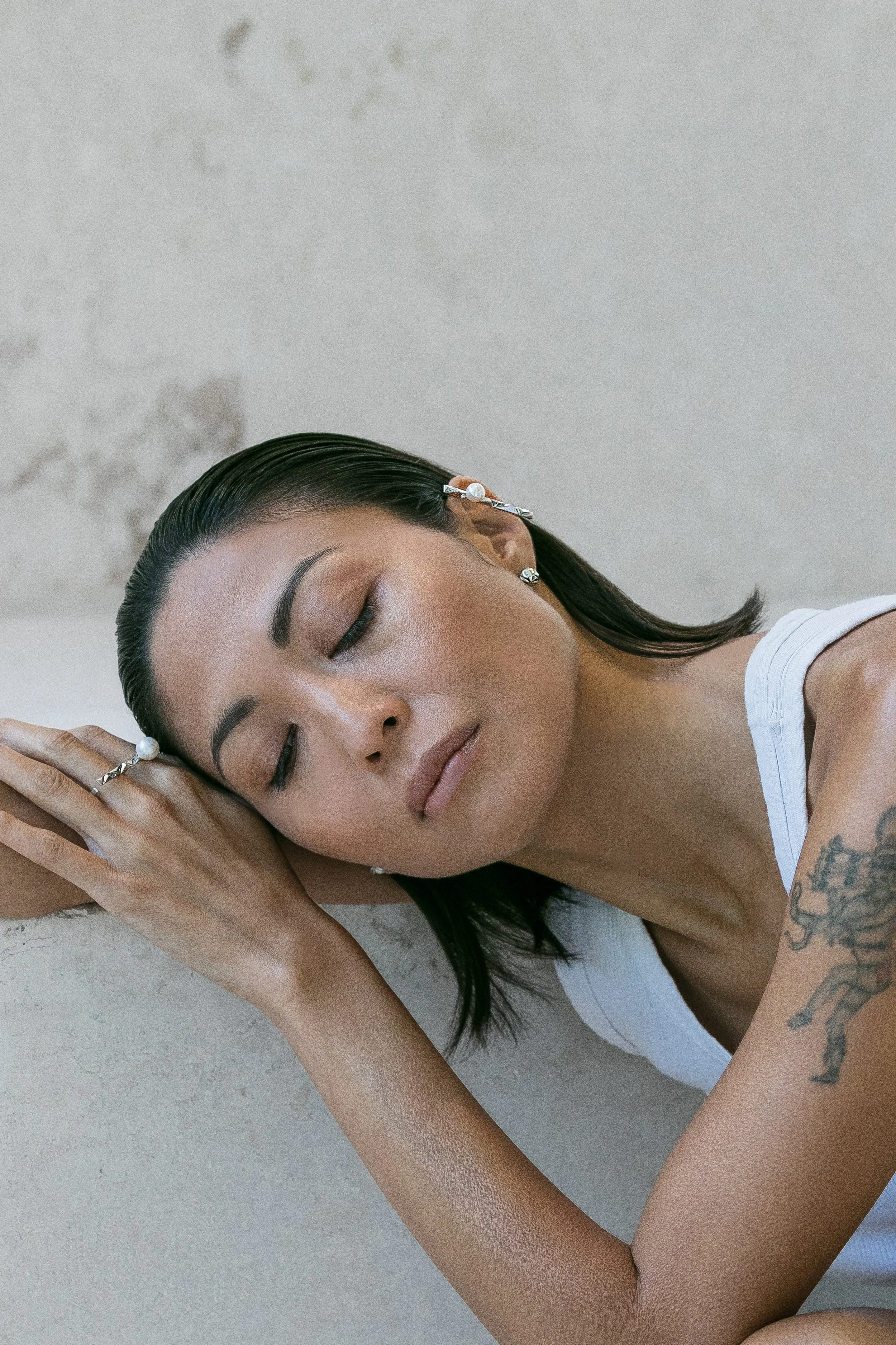 A woman with dark hair and makeup lying down with her eyes closed, resting her head on her arm, wearing pearl jewelry and a white top, with a tattoo on her upper arm.