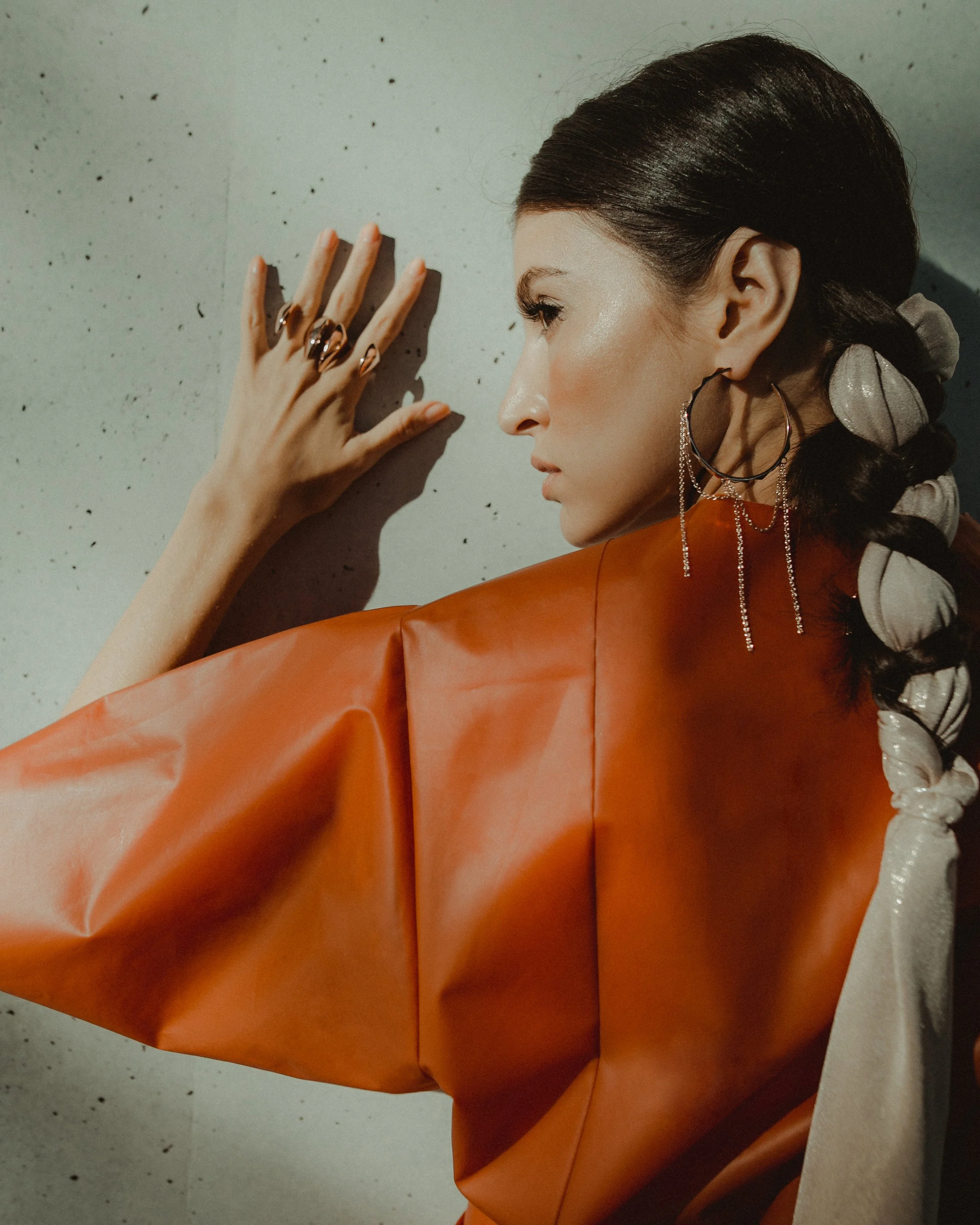 A woman with dark hair styled in a braided ponytail with gray hair ties, wearing large hoop earrings with chains, an orange jacket with puffed sleeves, and a silver satin ribbon in her hair, posing against a speckled wall.