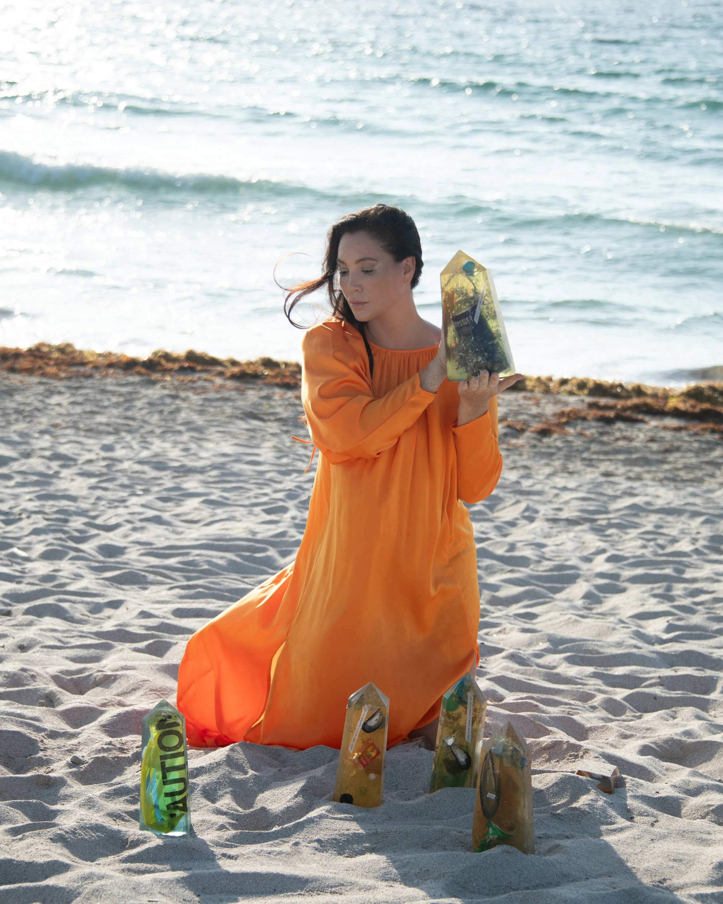 A woman in an orange dress kneeling on the beach, surrounding herself with used plastic bottles, some labeled caution, as she holds a plastic bottle close to her face during daytime.
