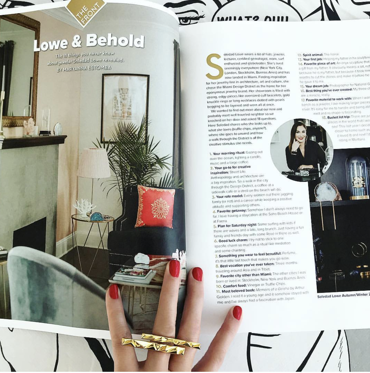 Open magazine featuring an article titled 'Lowe & Behold' with a photograph of a cozy living room corner, including a black armchair with a red pillow, a small table with books and decor, a tall floor lamp, and a potted plant. There's a mirror on the