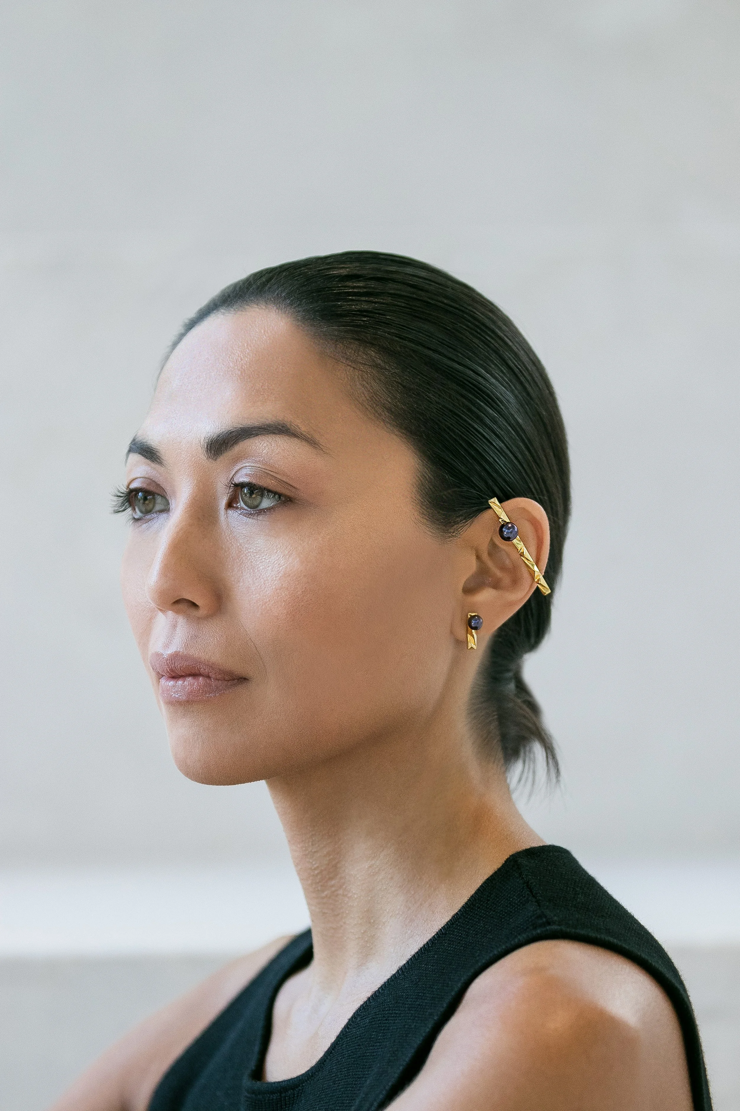 Close-up of a woman with slicked-back dark hair, wearing gold earrings with navy blue stones, dressed in a black sleeveless top, looking thoughtfully to the side.