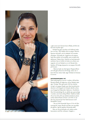 A woman with dark hair tied back, wearing a blue top and jewelry, sitting with her chin resting on her hand in an indoor setting.
