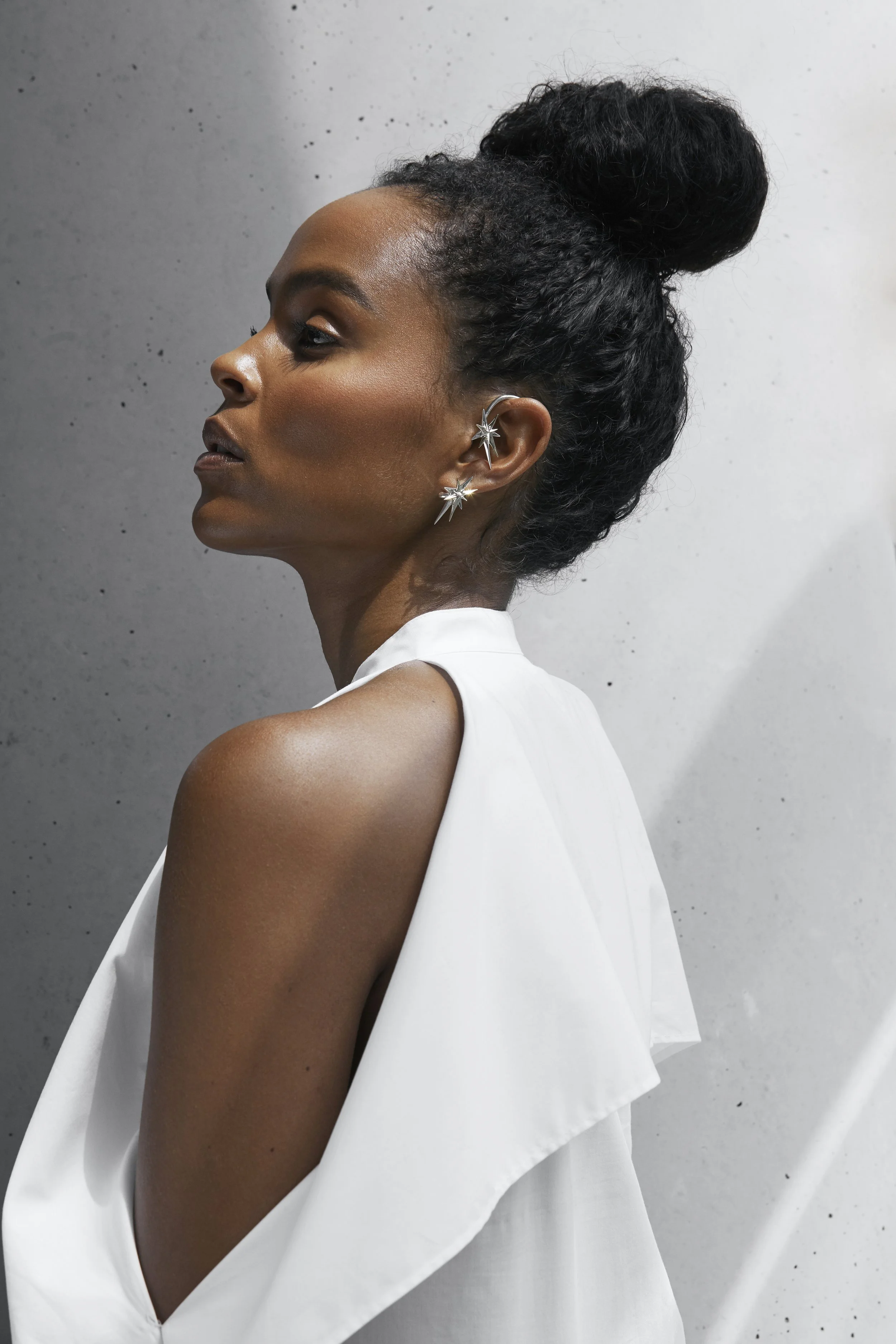 A side profile of a woman with dark hair styled in an updo, wearing star-shaped earrings and a white sleeveless dress, standing against a textured gray wall.