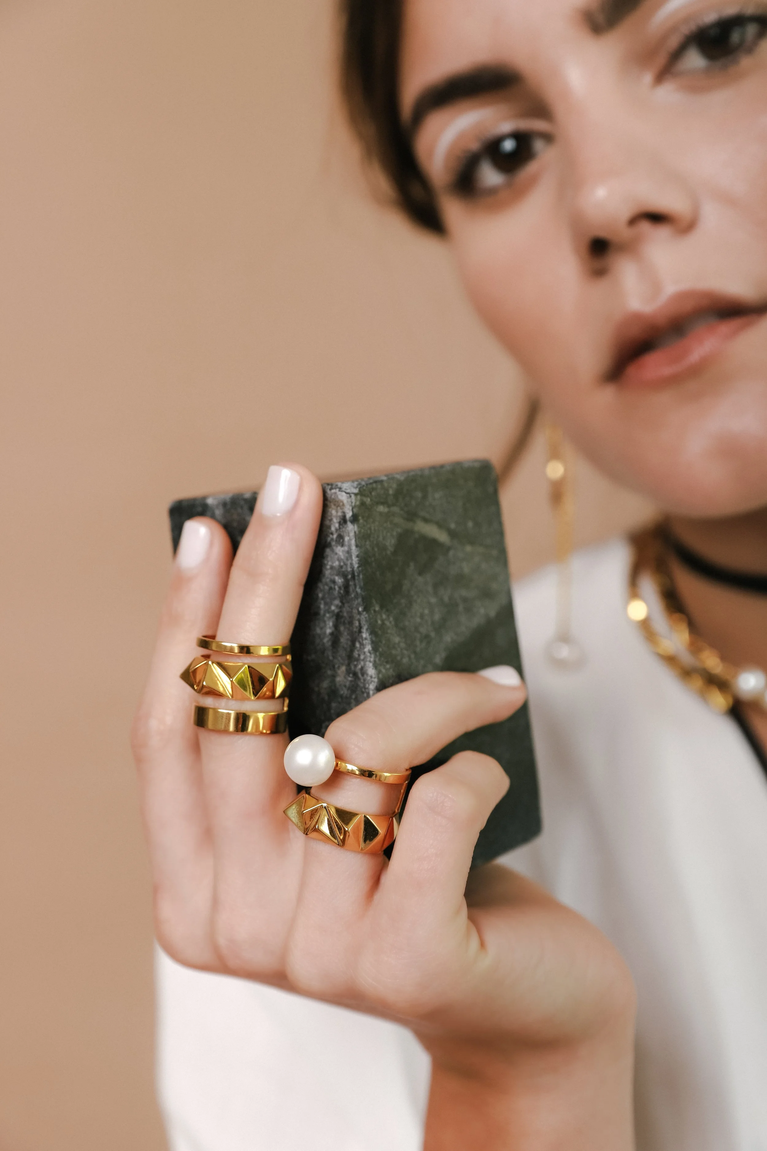 A woman holding a dark green stone, wearing multiple gold rings including one with a large white pearl, and gold jewelry.