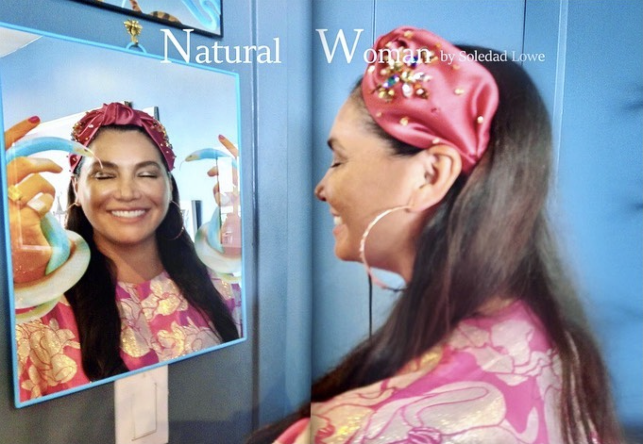 A woman with long dark hair, a pink headscarf with decorations, and hoop earrings is smiling and looking at her reflection in a mirror with a blue frame. She is wearing a pink patterned shirt. The mirror reflects her face and upper body, and the text