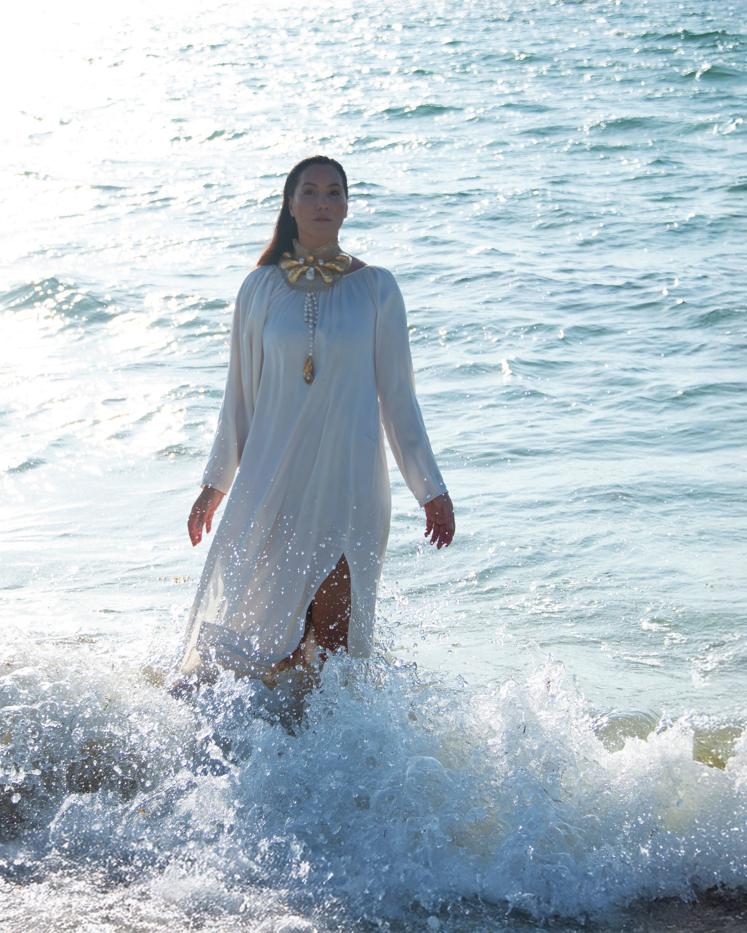 Woman in a white dress standing in the ocean waves near the shore, backlit by sunlight.