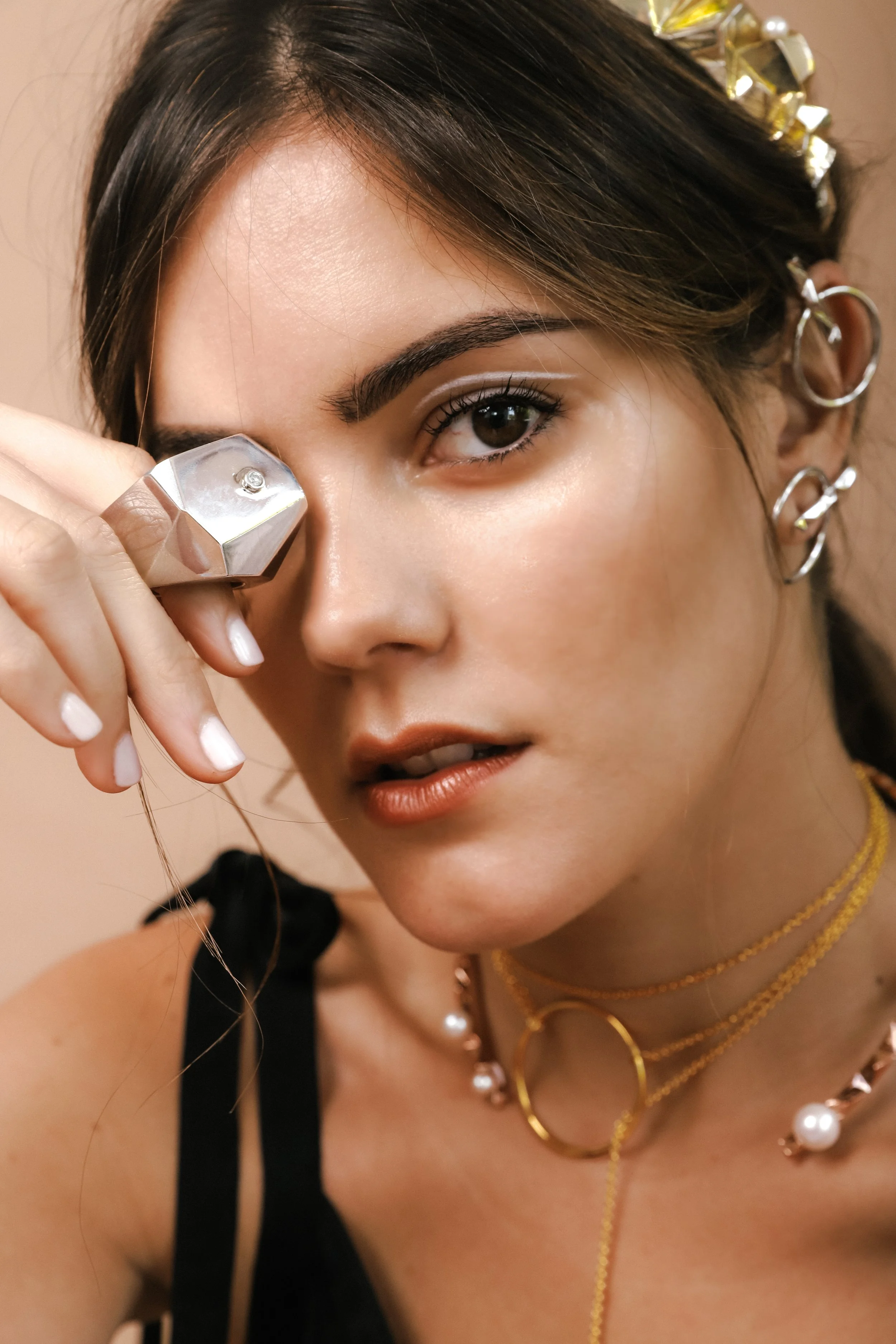 A woman with dark brown hair, styled with multiple earrings and a gold hair accessory, is holding a beauty device to her eye, with her face turned slightly to her right. She is wearing layered gold necklaces, a black top, and has makeup with defined 