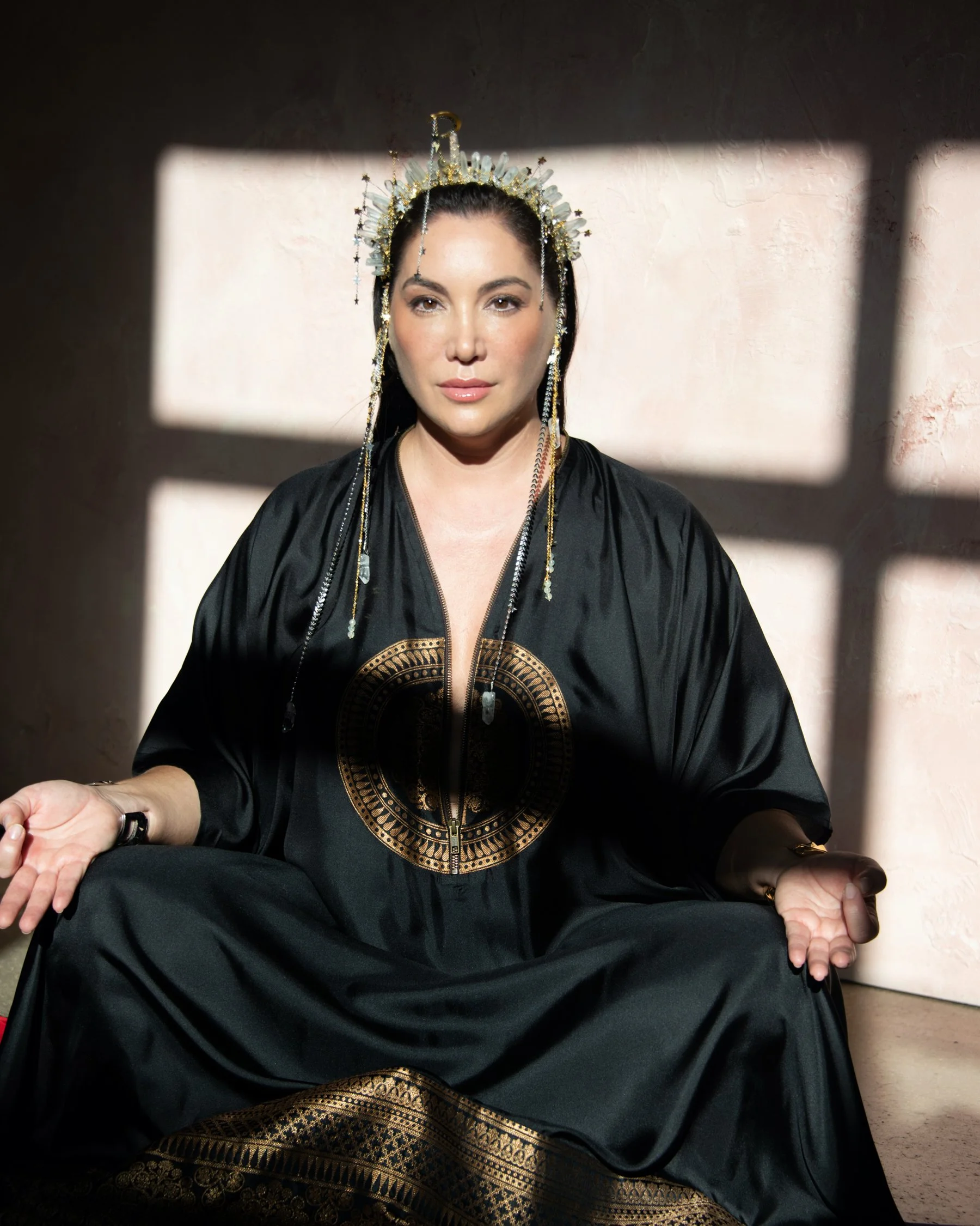 A woman with dark hair wearing a black robe with gold accents, sitting cross-legged with hands open, in front of a shadowed window that creates rectangular shadows on the wall behind her, wearing a headpiece with gold and silver details.
