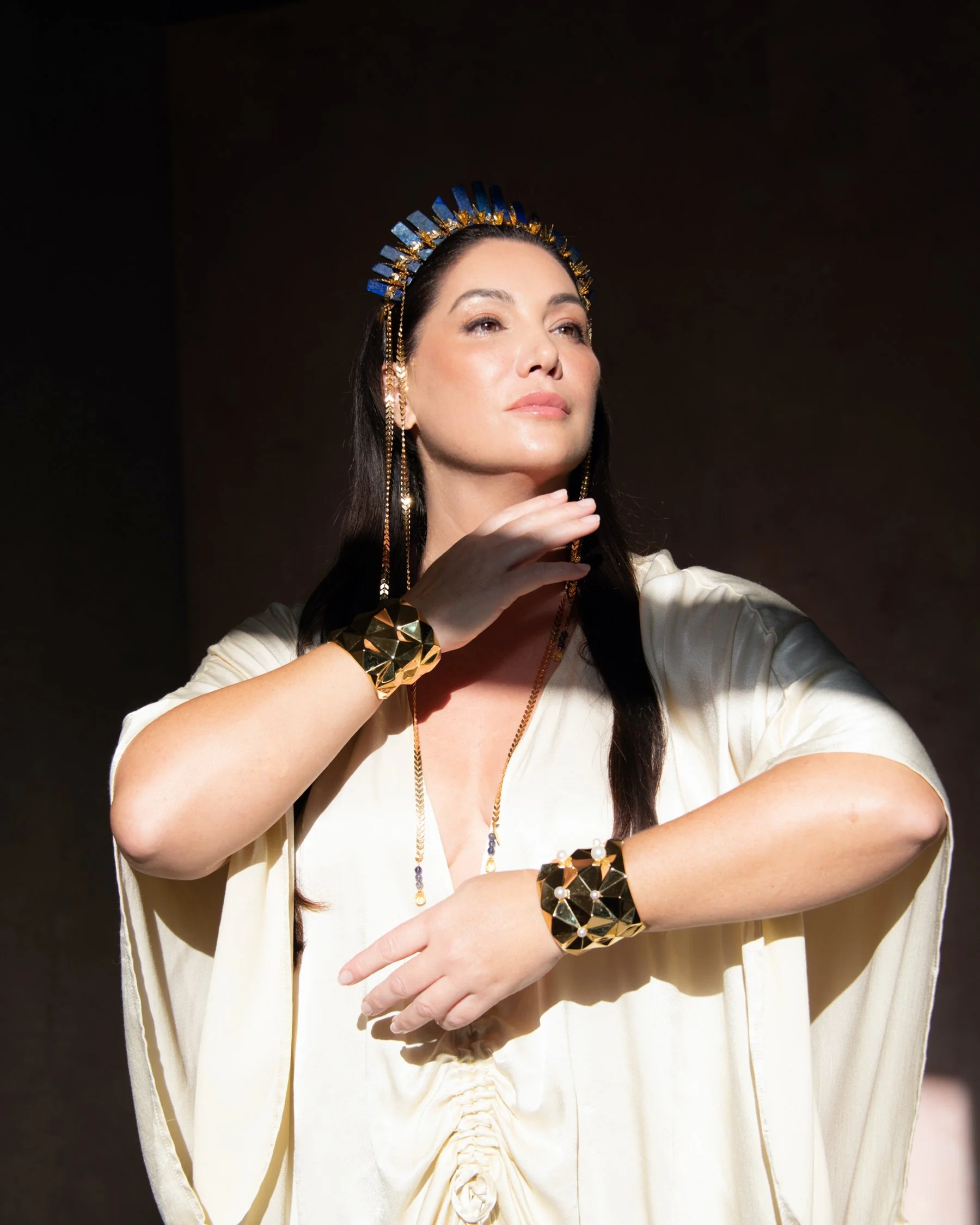 A woman with long dark hair wearing a cream-colored dress, gold jewelry including a crown-style headpiece, large bracelets, and a necklace, posing with her hand near her face against a dark background.