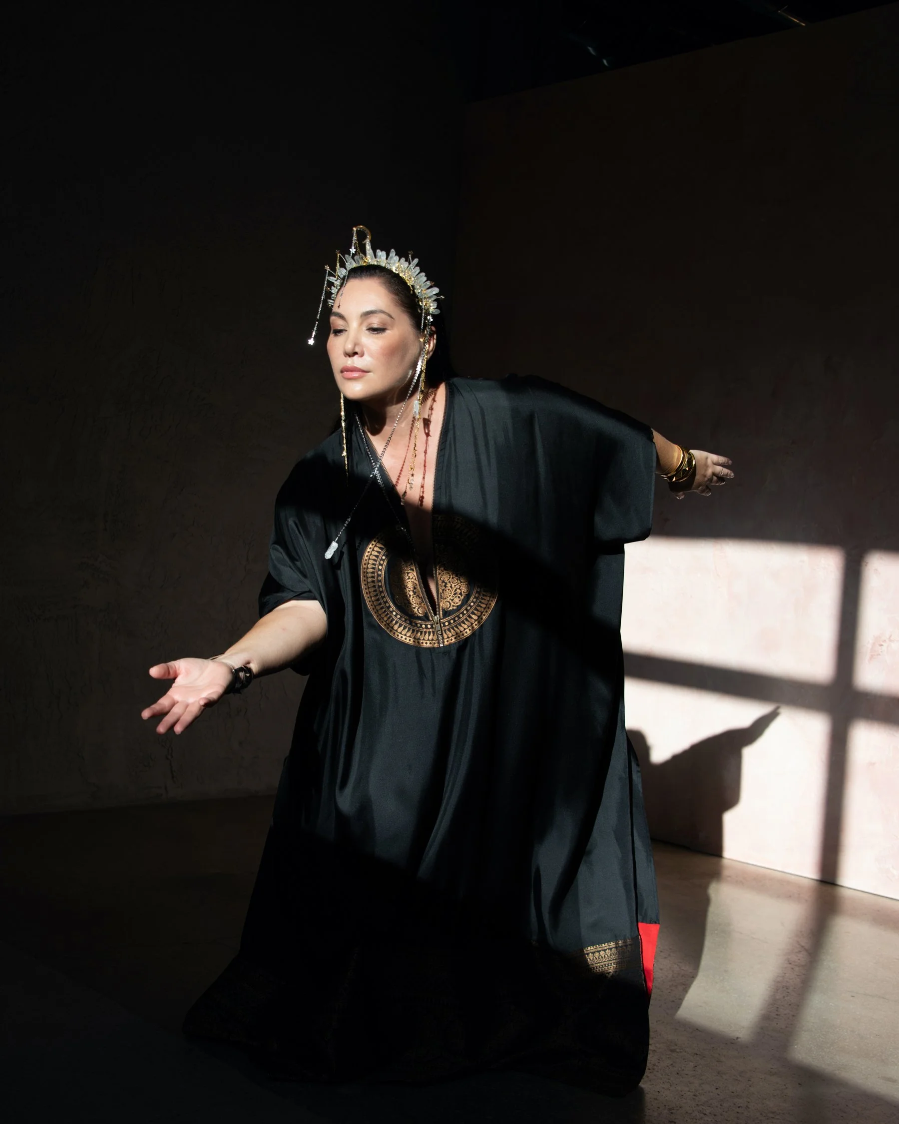 A woman wearing a black dress with a gold pattern, gold jewelry, and a decorative silver headdress with spikes posing in front of a plain wall, with sunlight creating a grid shadow on the wall.