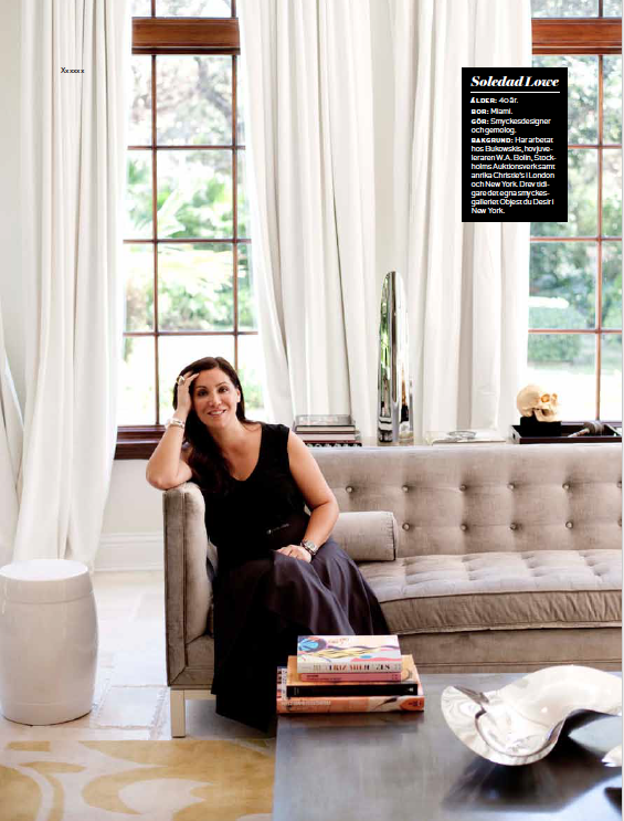 Woman sitting on a beige sofa in a bright, modern living room with large windows and white curtains, with a stack of magazines on a coffee table