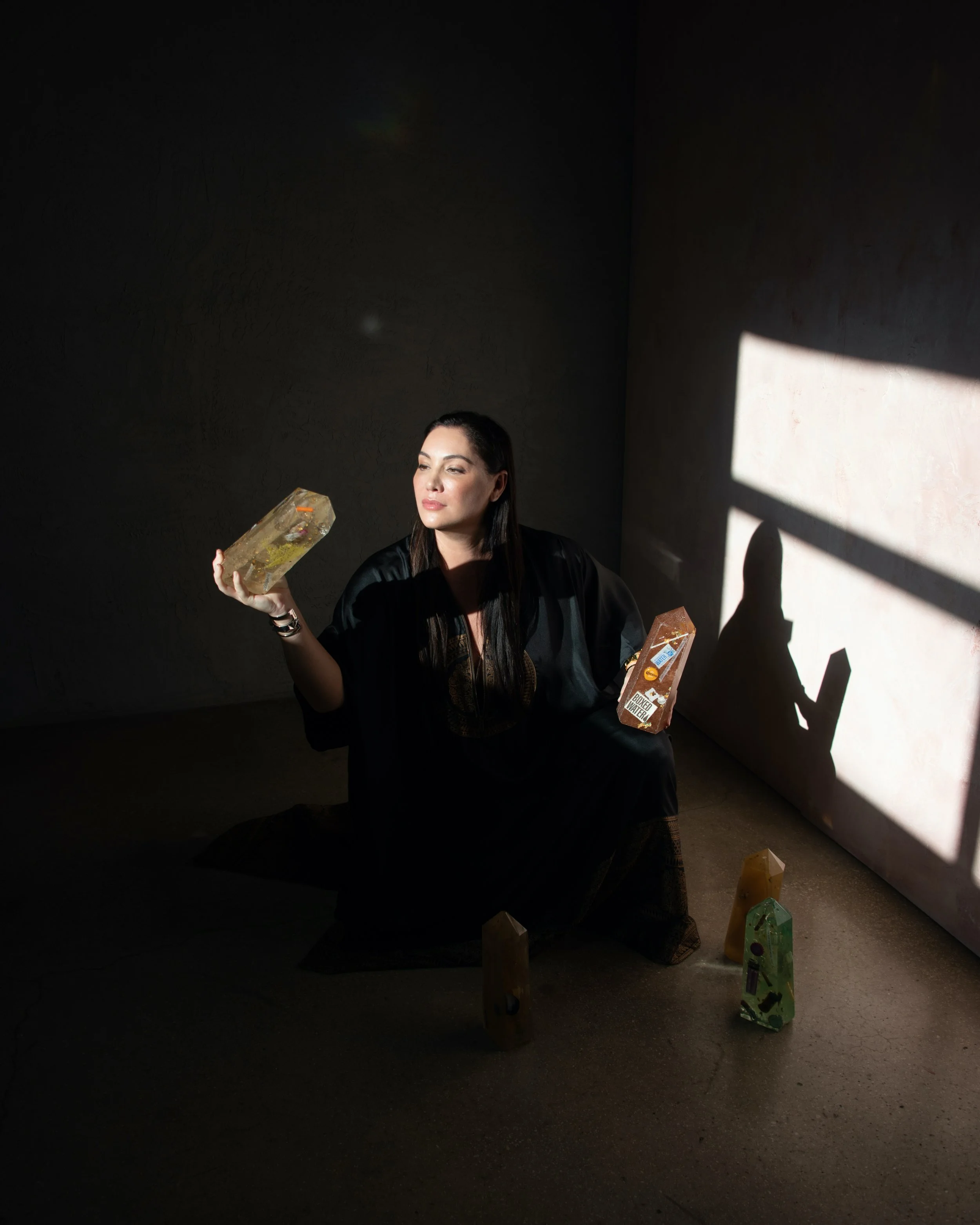Woman sitting on the floor in a dimly lit room, holding a large rectangular object resembling a soap bar in each hand, with three similar objects on the ground nearby and a shadow of her holding a book cast on the wall.