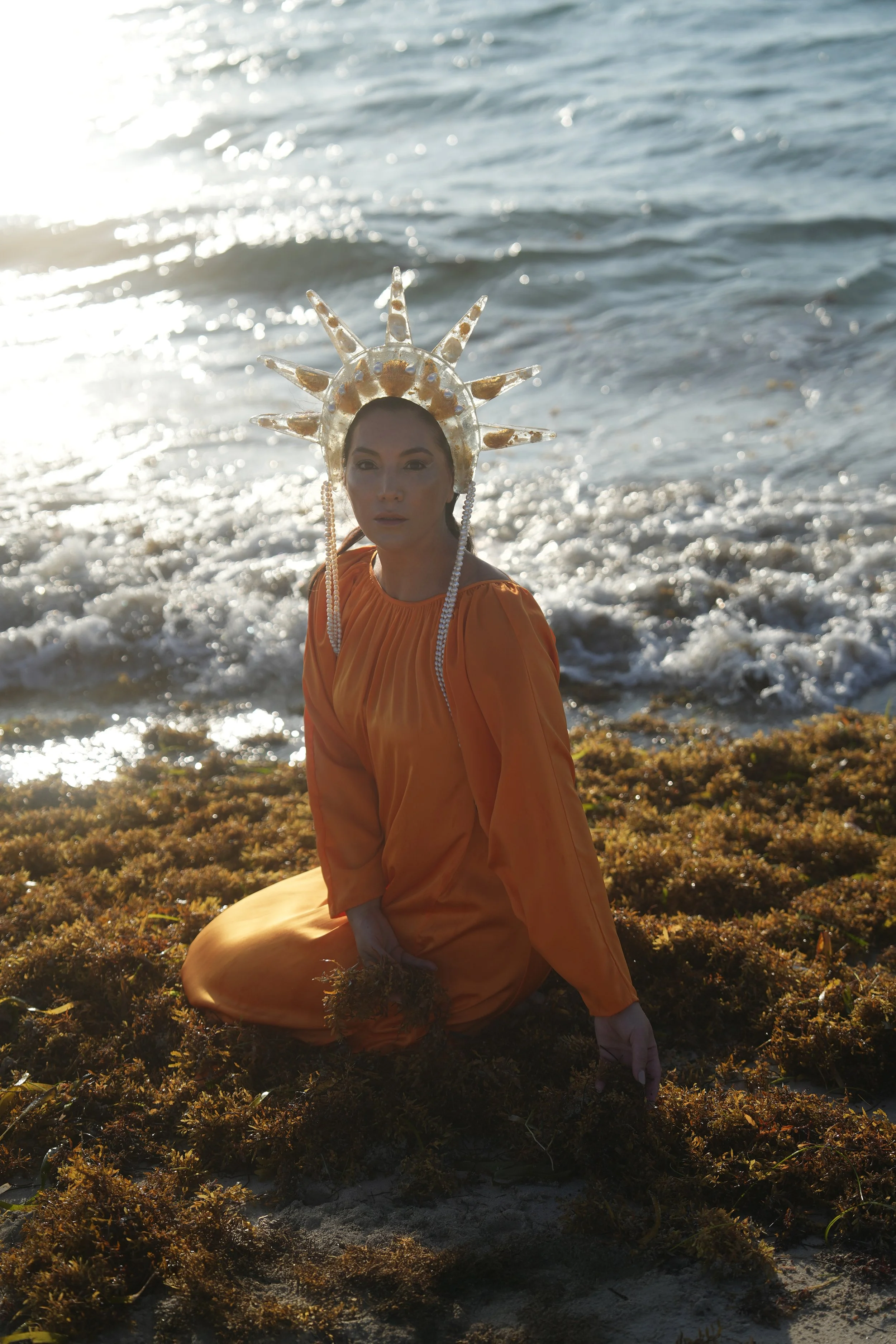 A woman in an orange dress kneeling on a beach with seaweed, wearing a large seashell crown, with ocean waves in the background.