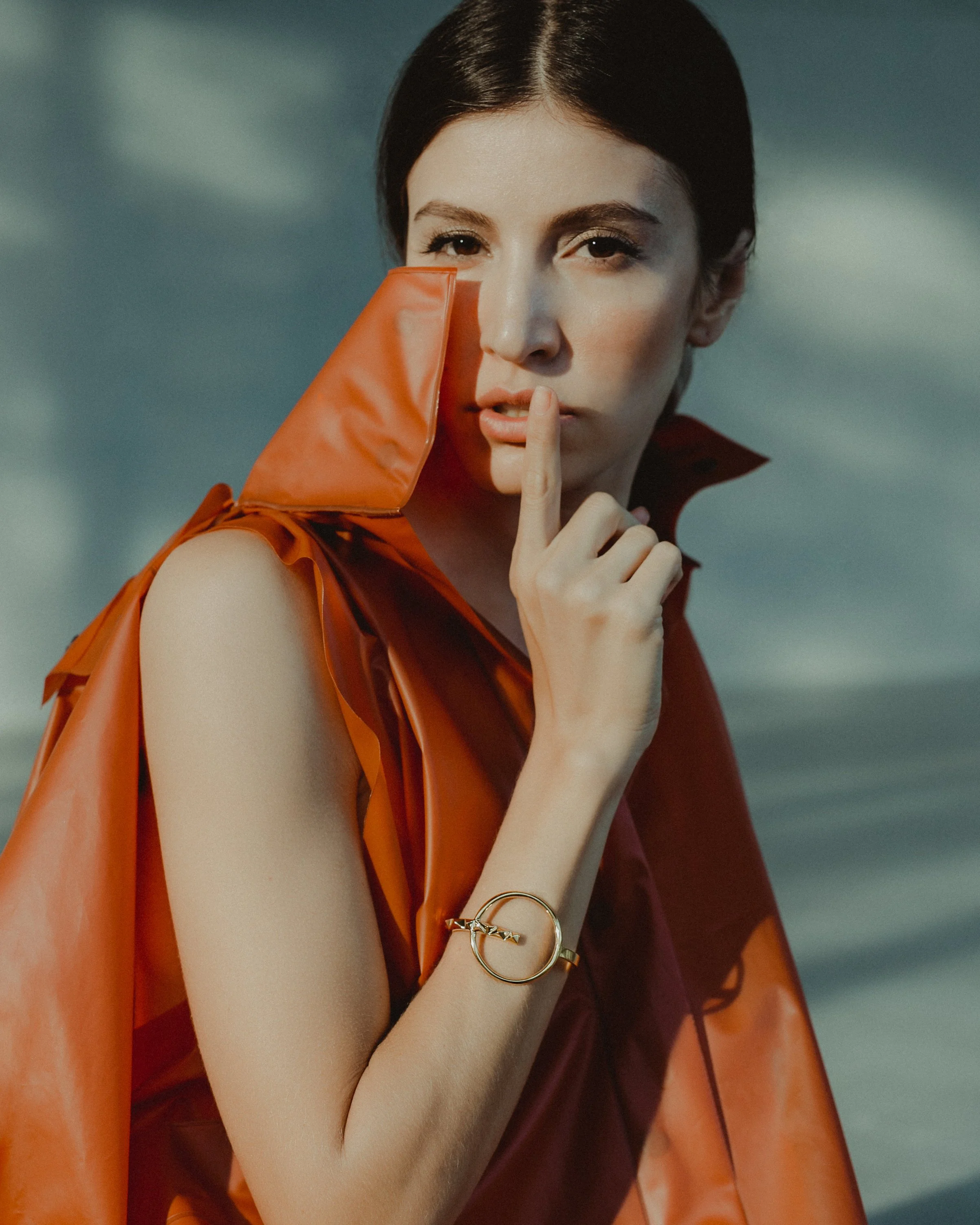 A woman with dark hair wearing an orange sleeveless outfit and a gold bracelet, holding her finger to her lips in a shushing gesture.