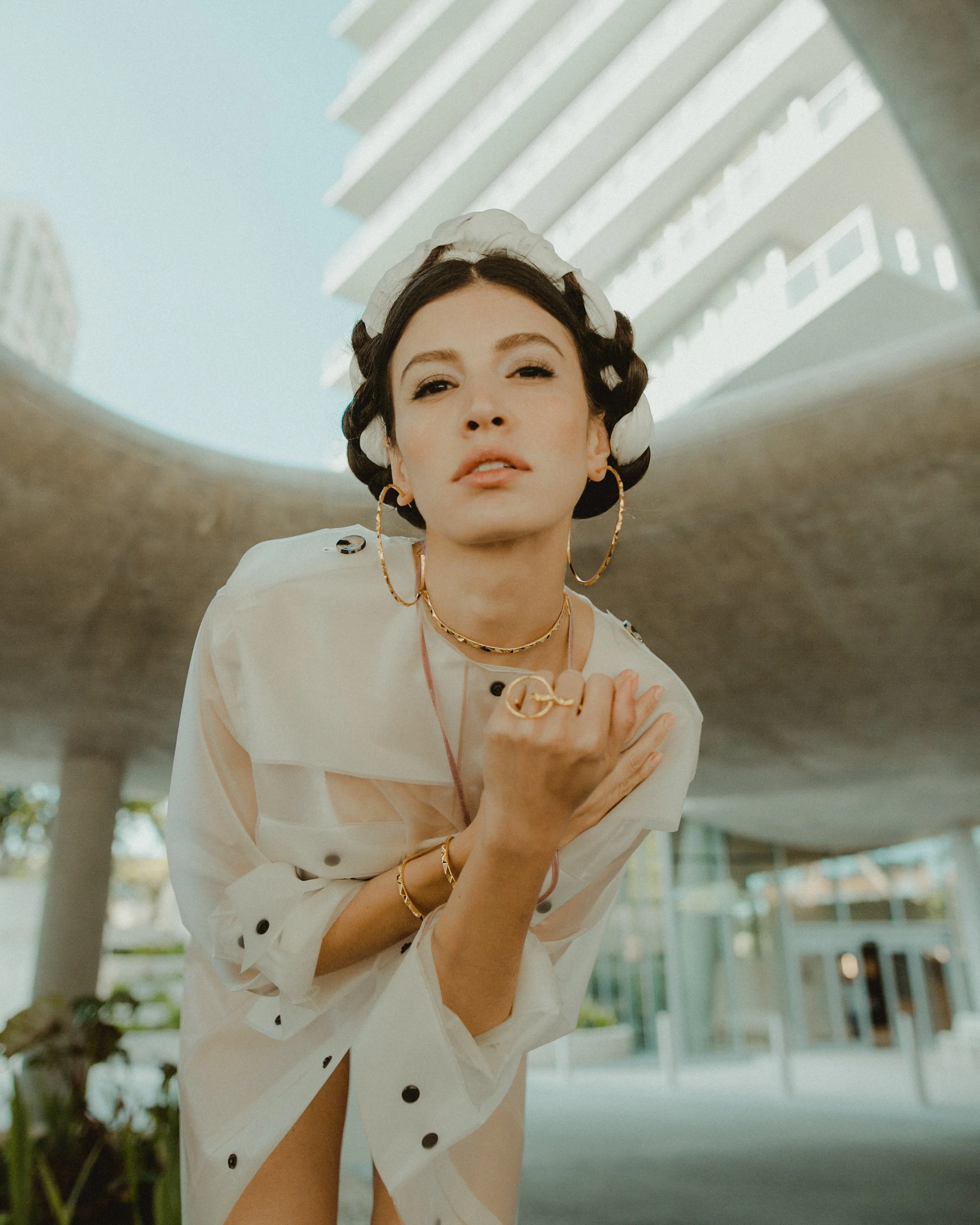 A woman with dark hair in curlers, wearing gold hoop earrings and jewelry, posing outdoors with modern building structures in the background.