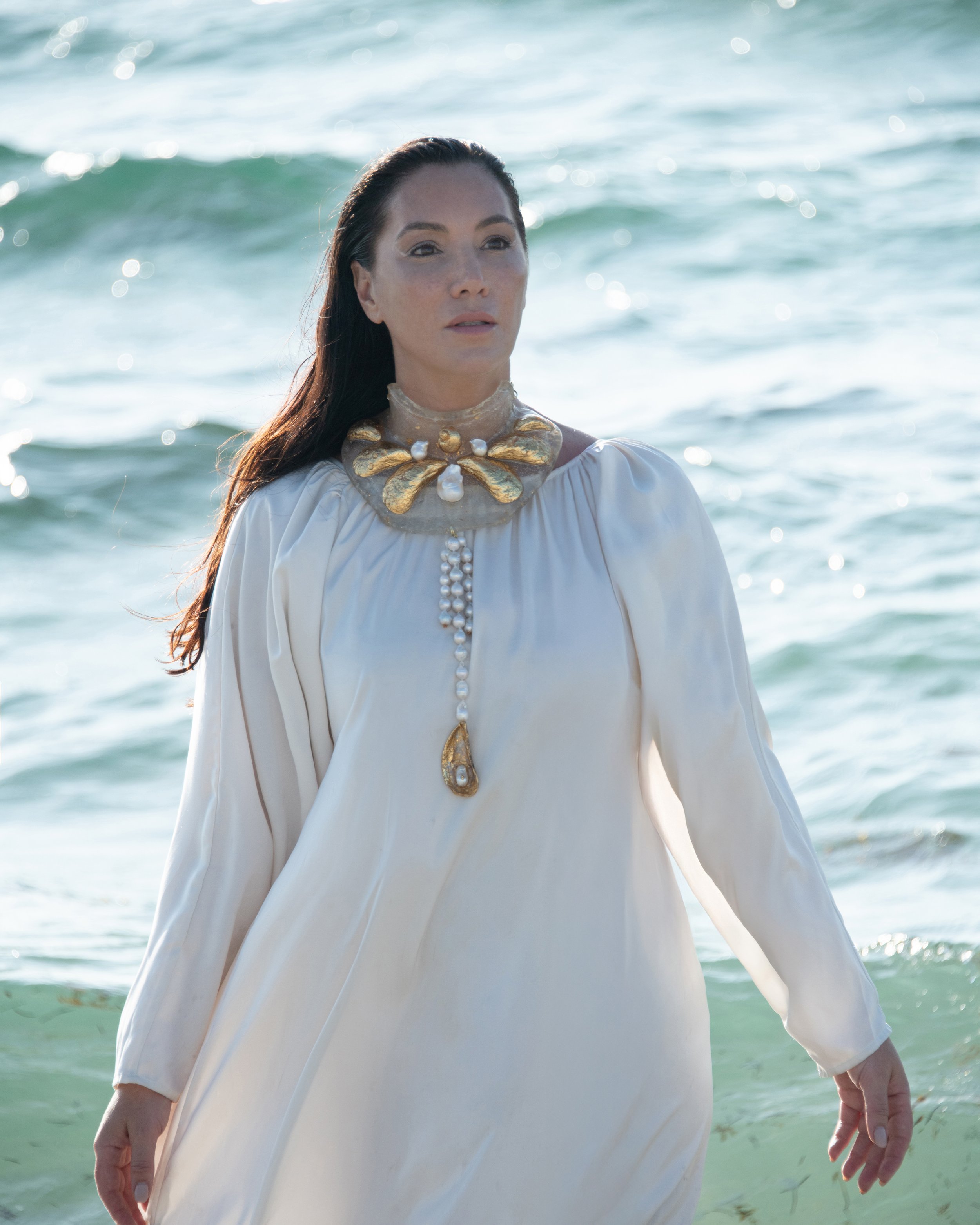 A woman with dark hair walking out of the ocean, wearing a white dress with an elaborate gold and pearl necklace or collar.