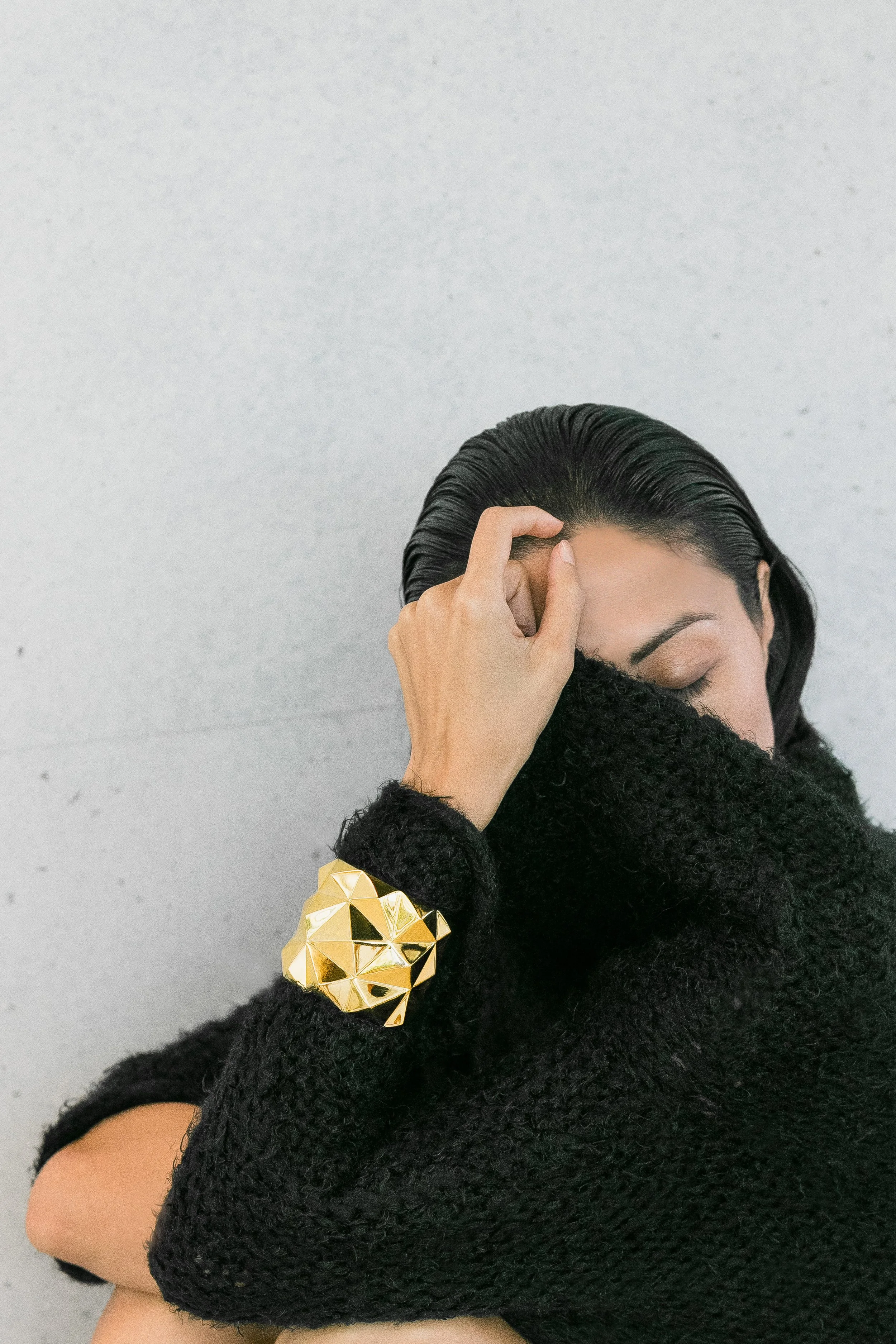 A woman with her eyes closed, wearing a black sweater and a gold geometric bracelet, touching her forehead with her right hand against a gray wall background.