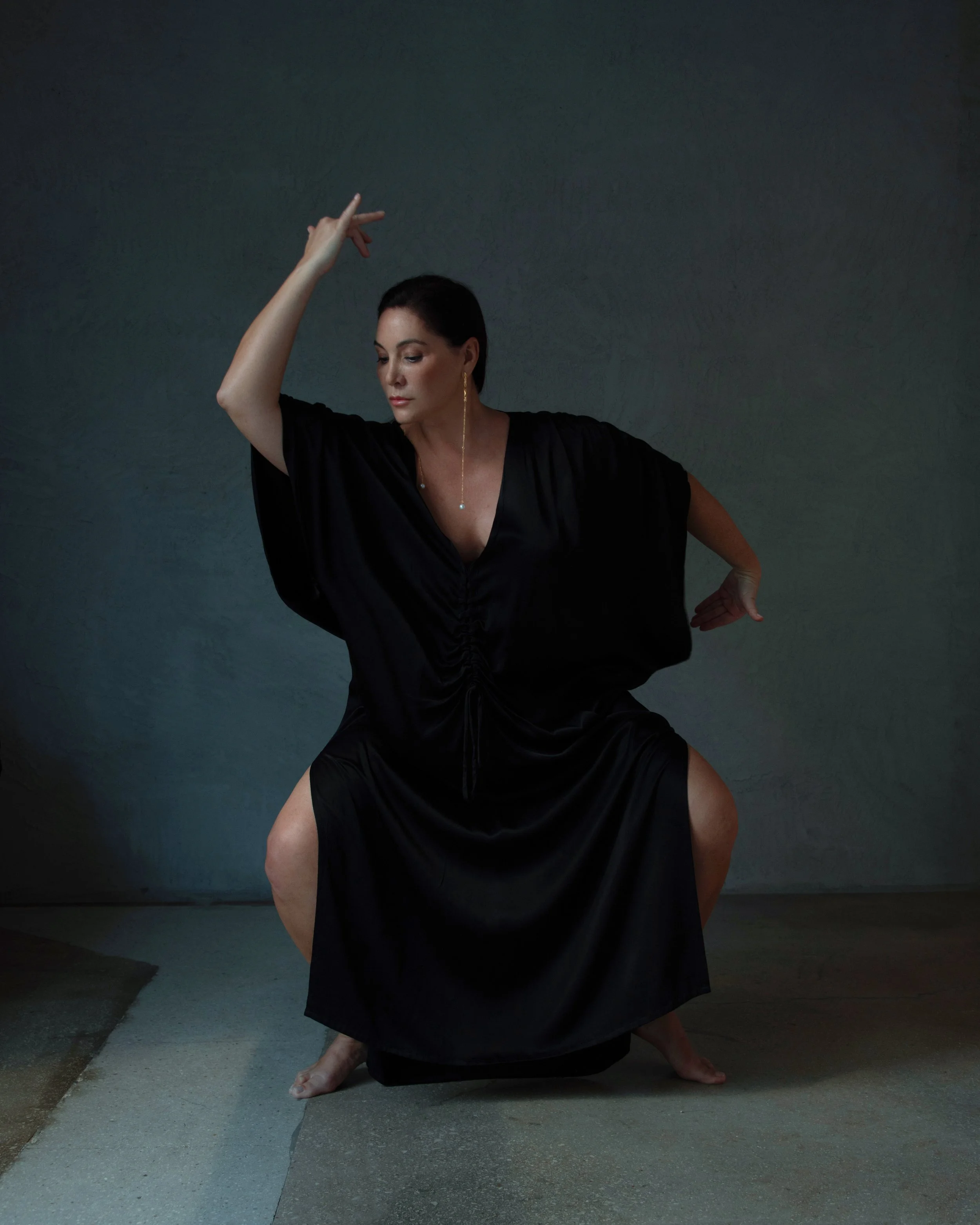 A woman in a black dress is performing a dance pose on a gray concrete floor against a dark textured wall.