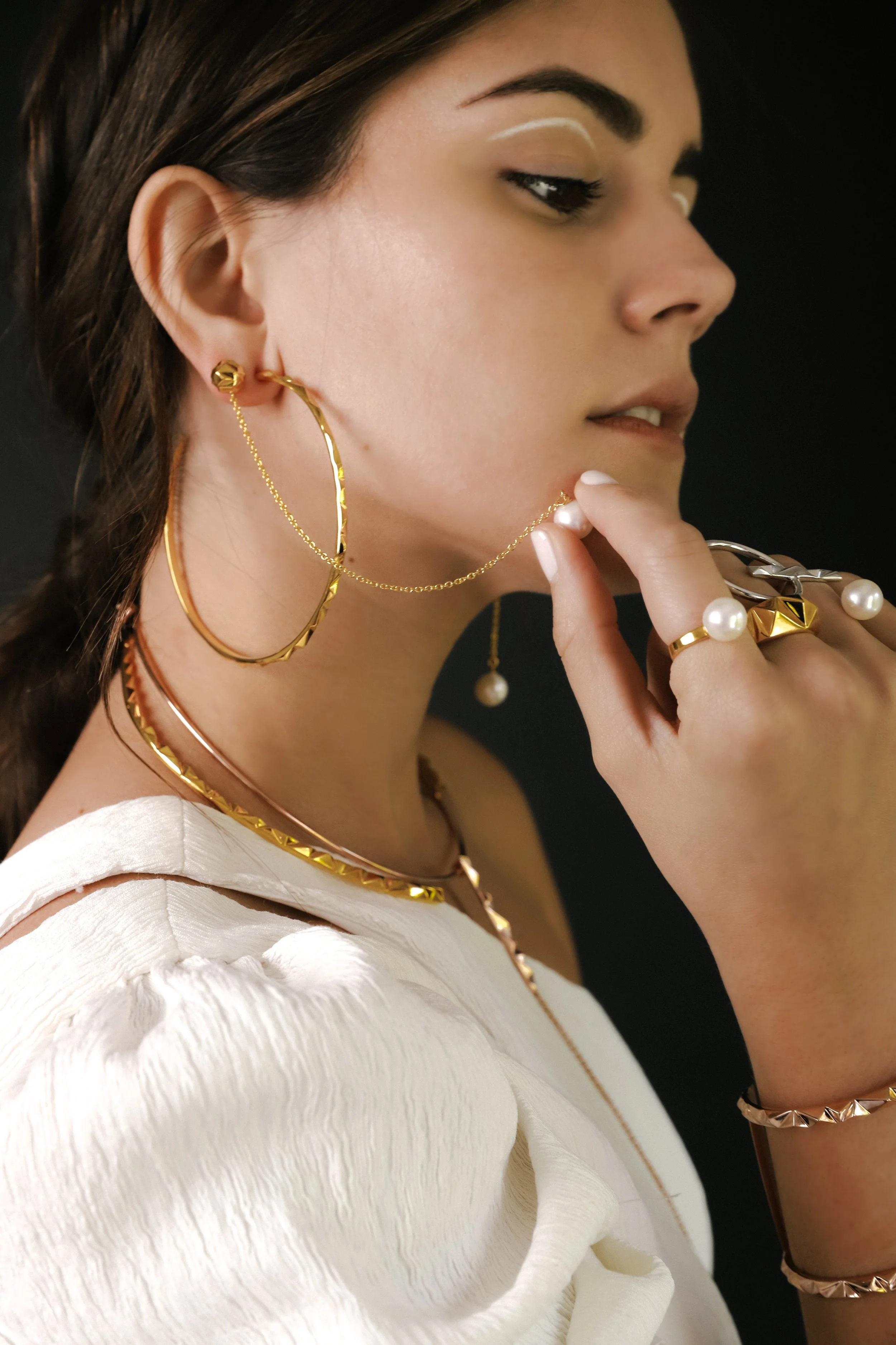 Close-up of a woman with dark hair wearing gold jewelry, including earrings, necklaces, rings, and bracelets, against a dark background.
