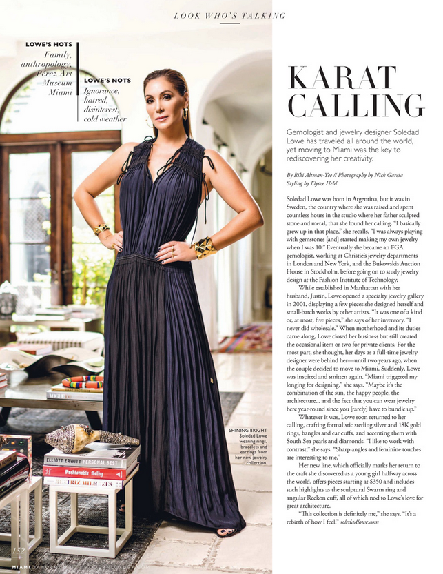 A woman wearing a navy blue pleated dress standing in a bright room with a white background, next to a gaming table and a stack of books.