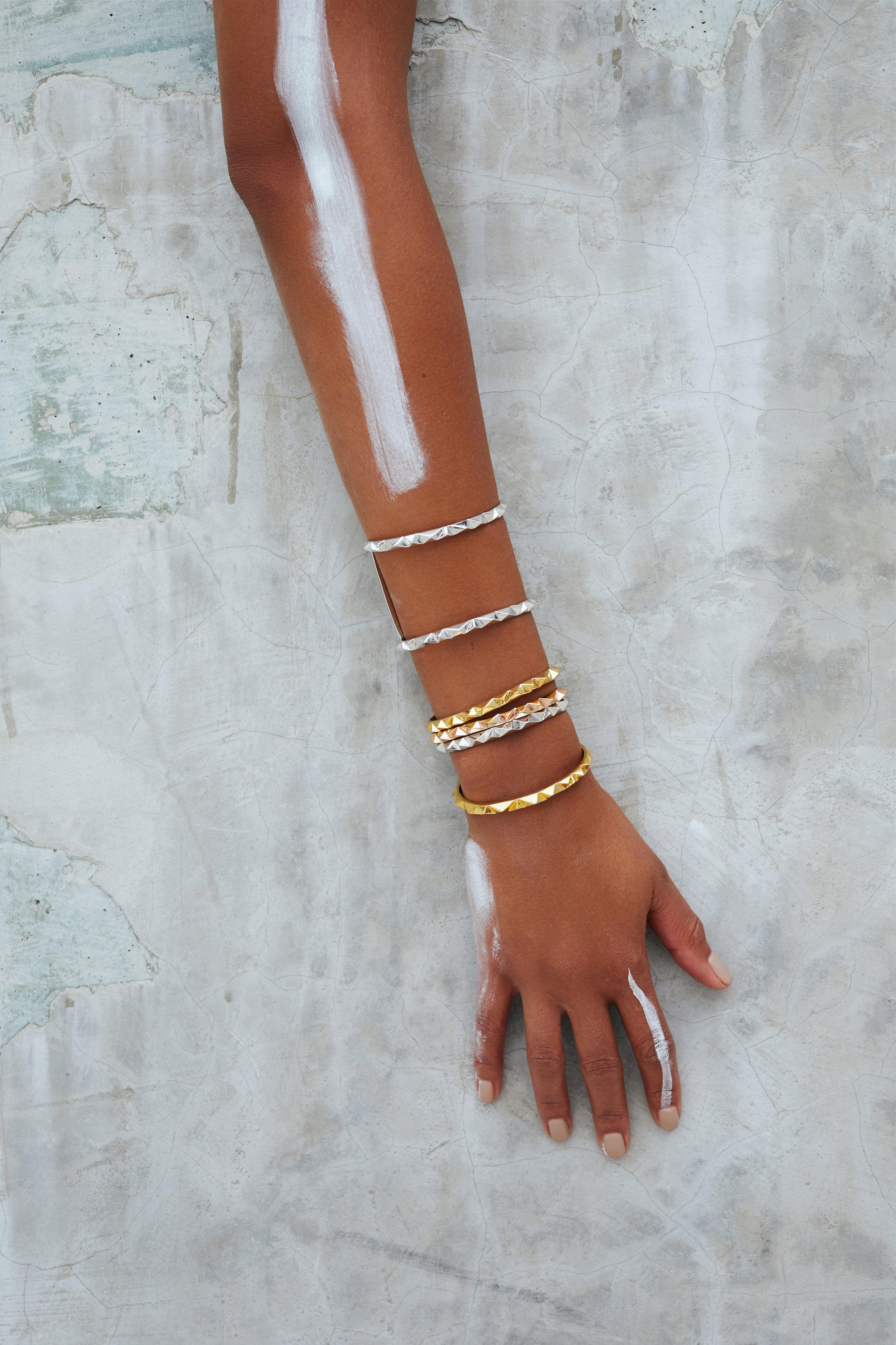 A person's arm and hand resting on a textured, painted concrete surface. The arm is adorned with multiple silver and gold studded bracelets, and there is white paint or chalk on the arm and hand. The background is a rustic, weathered wall.