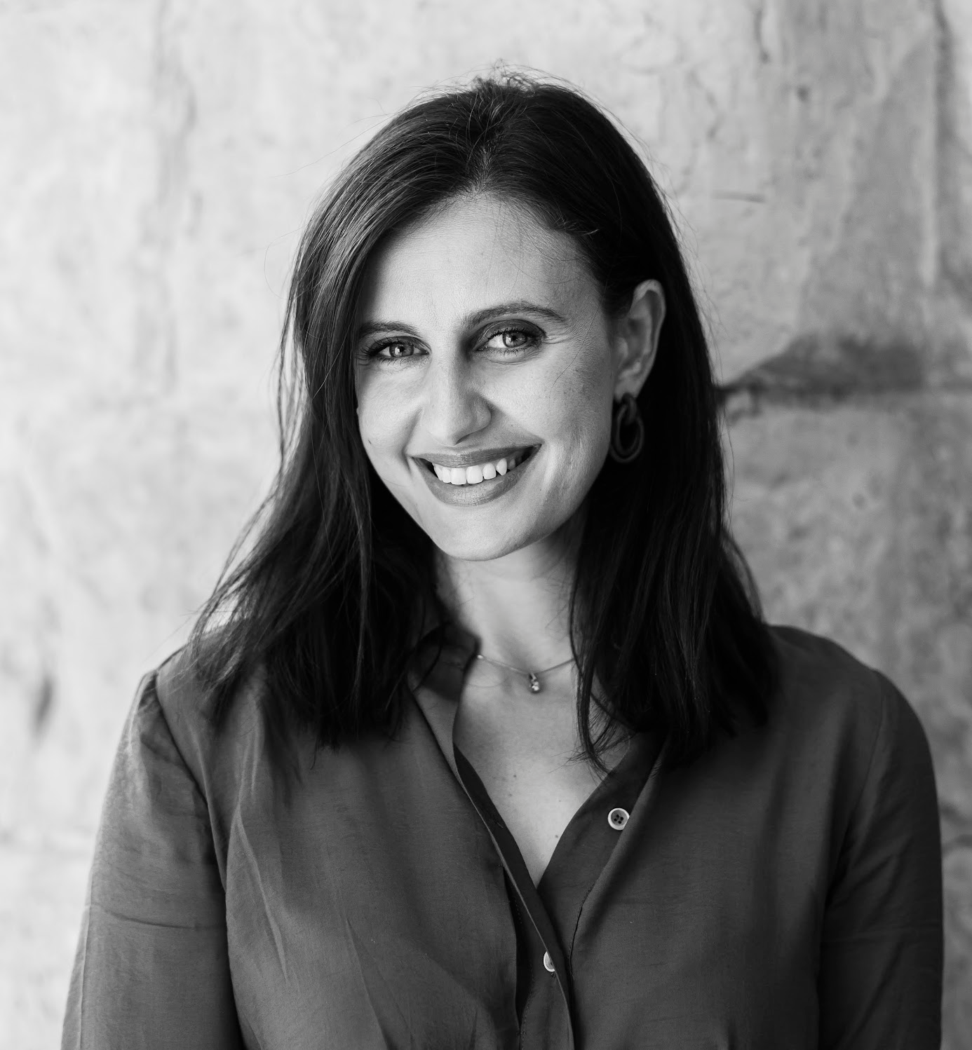 A smiling woman with dark hair and earrings, wearing a button-up shirt, standing in front of a textured wall.