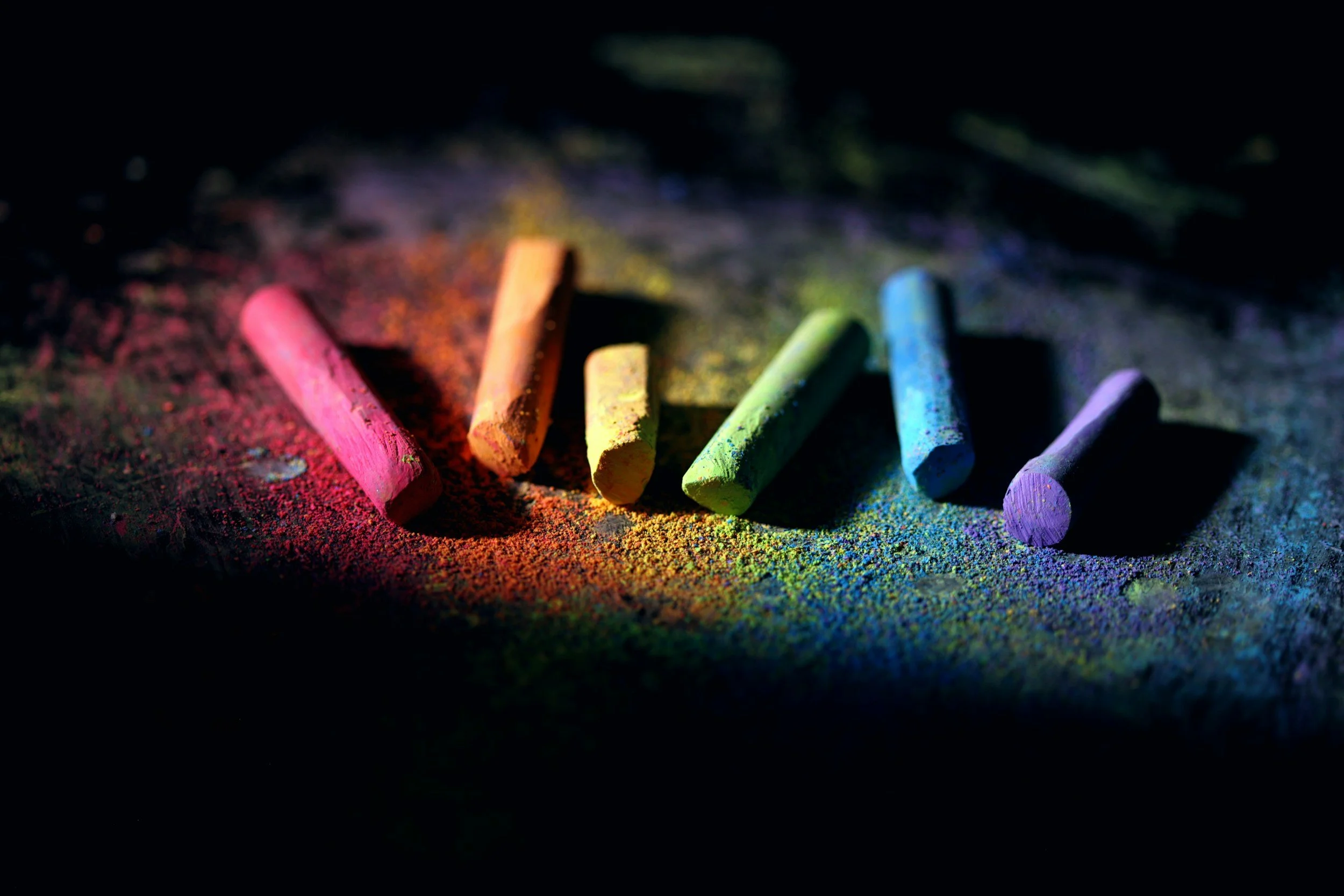 Colorful chalk sticks on a dark surface with colorful chalk dust around them.