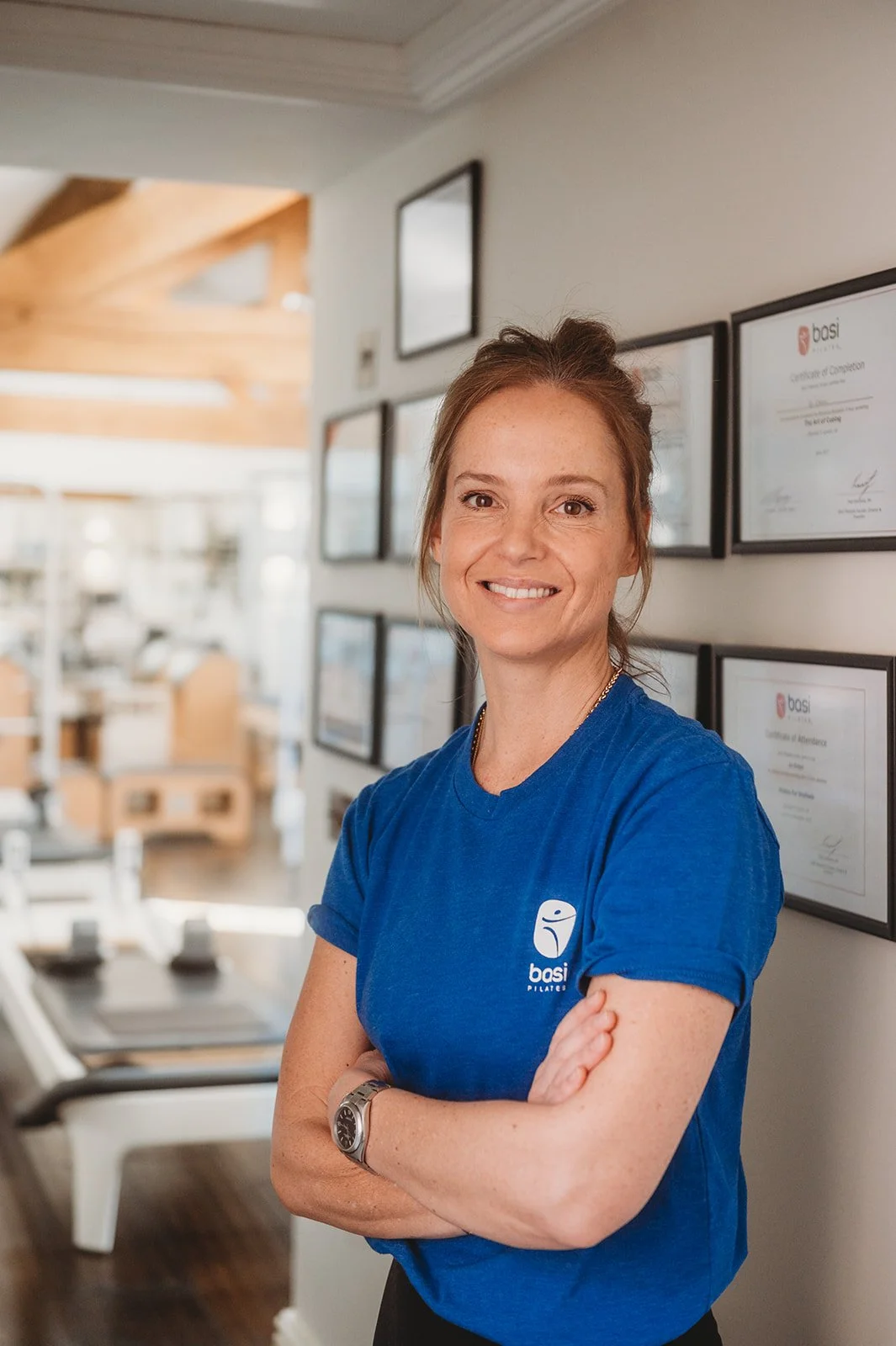 Female pilates teacher smiles with a BASI pilates T-Shirt on in front of a wall of certificates labeled with BASI pilates accomplishments.