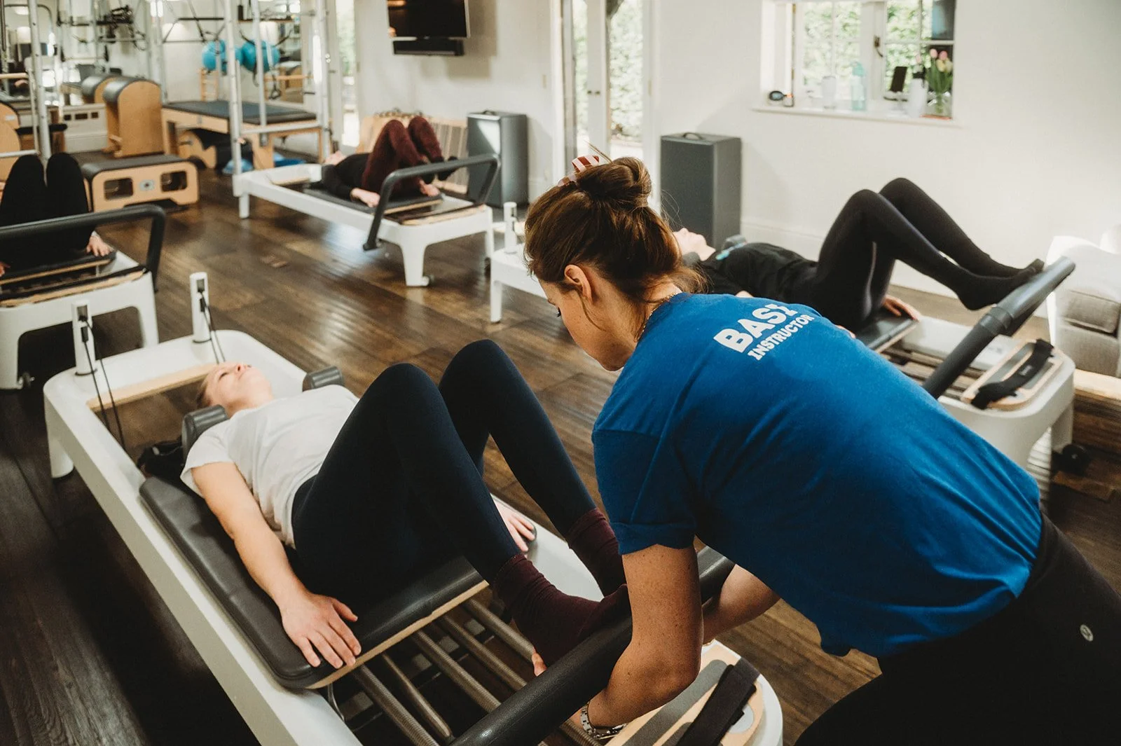 BASI trained pilates teacher corrects foot positioning of one of her students during a reformer pilates class while the four students la on their backs in the renovated barn on their reformers