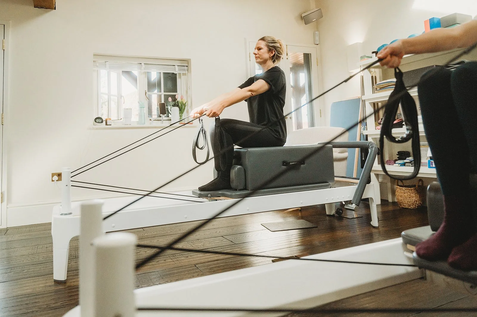 Two reformer pilates student sit on the edge of their reformers, pulling tautly on the cables