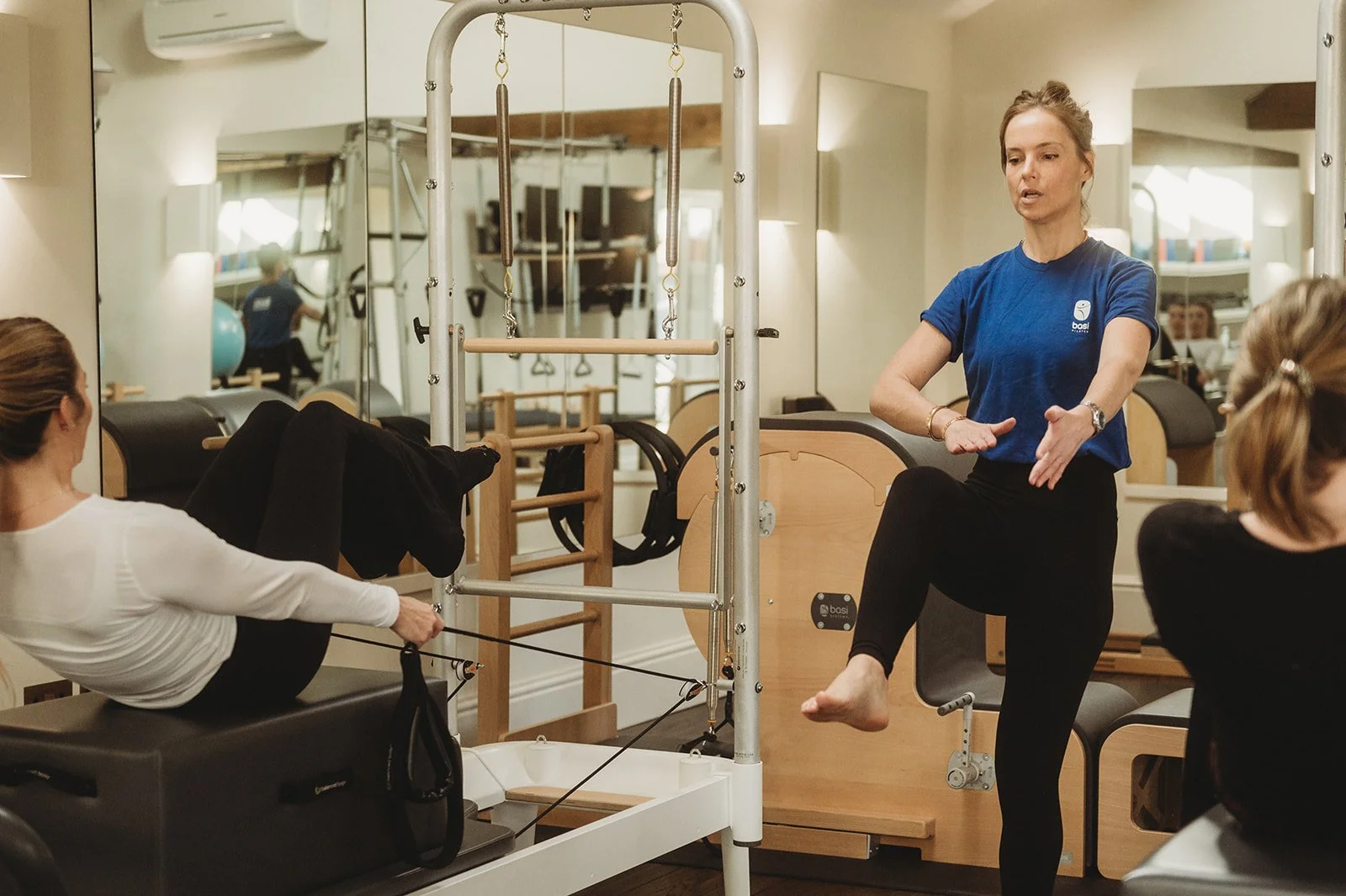 Reformer pilates teacher stands in front of the class and demonstrates the movement with her hands and legs 