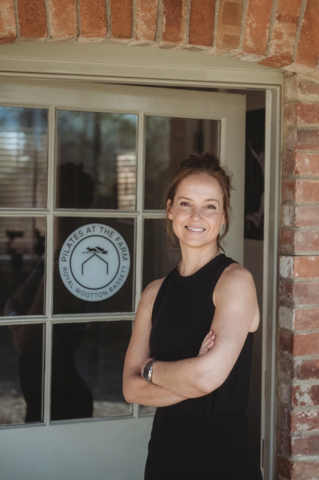 Smiling woman with brown hair standing in front of white door next to a logo reading 'Pilates at the Farm'