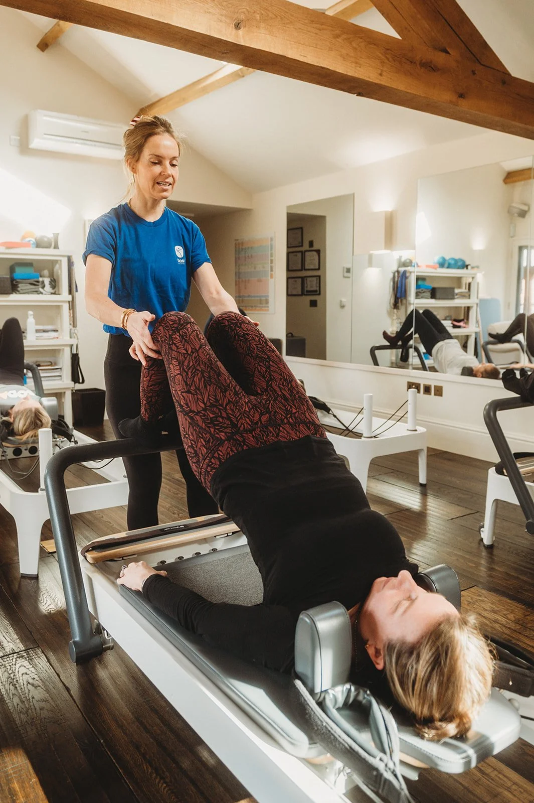 BASI pilates instructor lightly corrects the leg positioning of her student while her student is braced in the bridge position on the reformer