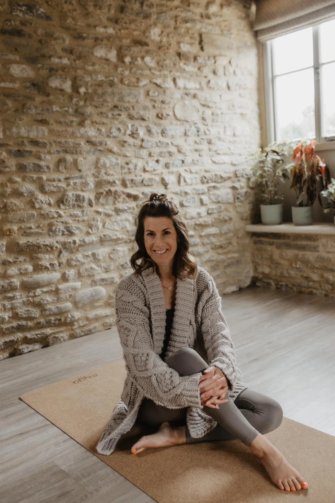 Female pilates teacher smiles with her brown hair half up half down as she sits comfortably on a mat in a thick beige cardigan