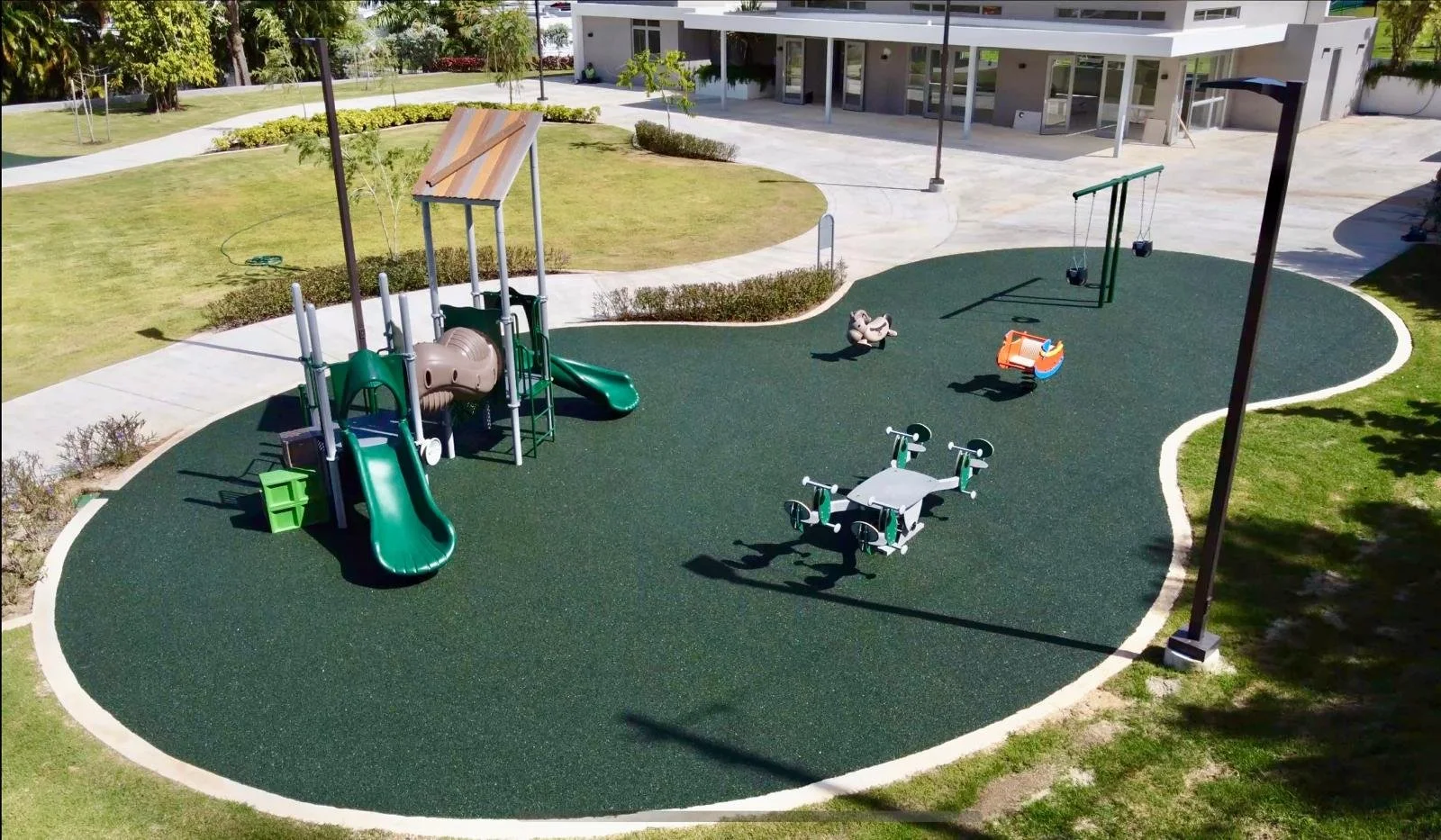 Playground with slides, swings, a spring rocker, a see-saw, and a roundabout on a rubber surface, surrounded by grass and bushes.