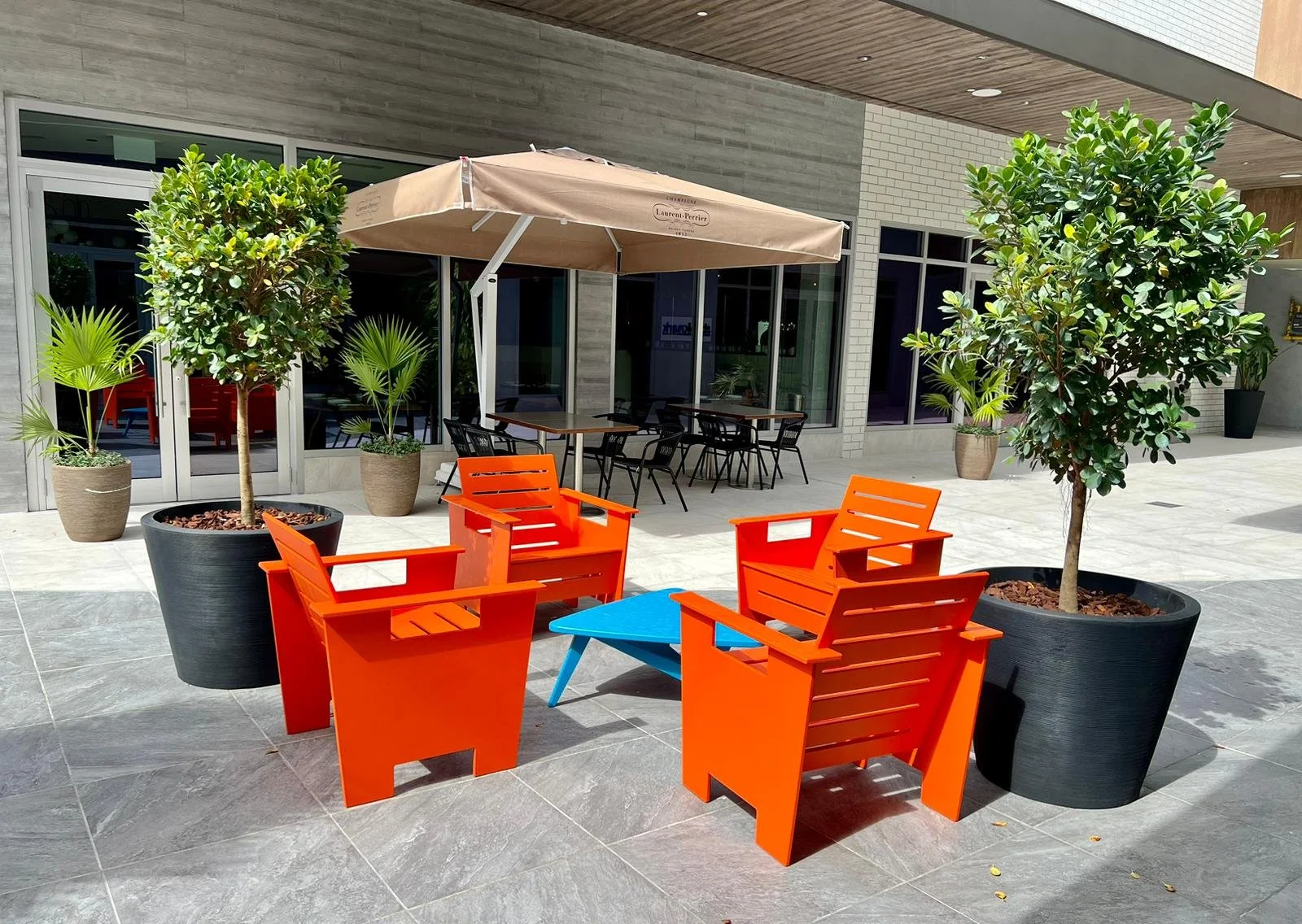 Outdoor seating area with two large potted trees, bright orange chairs, a small blue table, a large umbrella, and additional seating in the background, next to a building with large windows.
