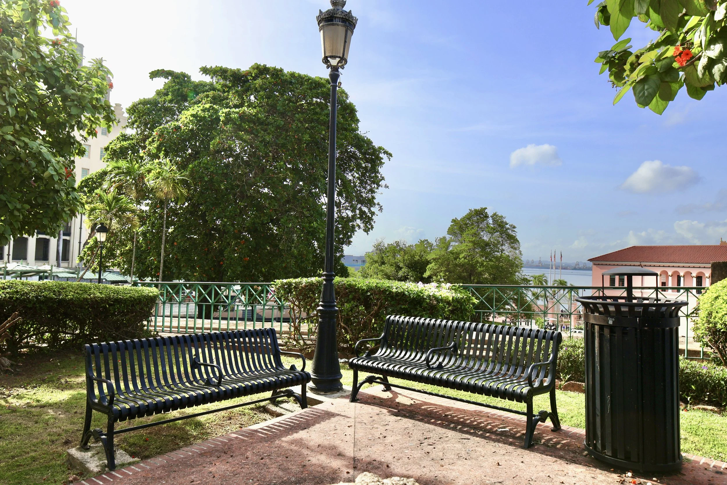 Public park with two black metal benches, a trash can, a street lamp, green bushes, and tall trees under a blue sky with some clouds. There is a pinkish building with flags in the background.
