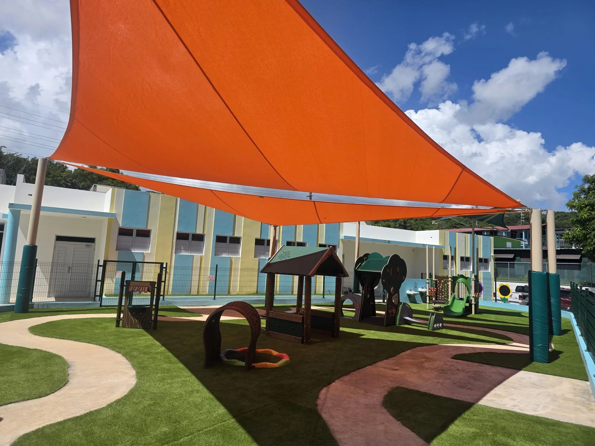 Children's outdoor playground with a big orange shade canopy, winding paths, playground equipment, and a colorful building in the background under a partly cloudy sky.