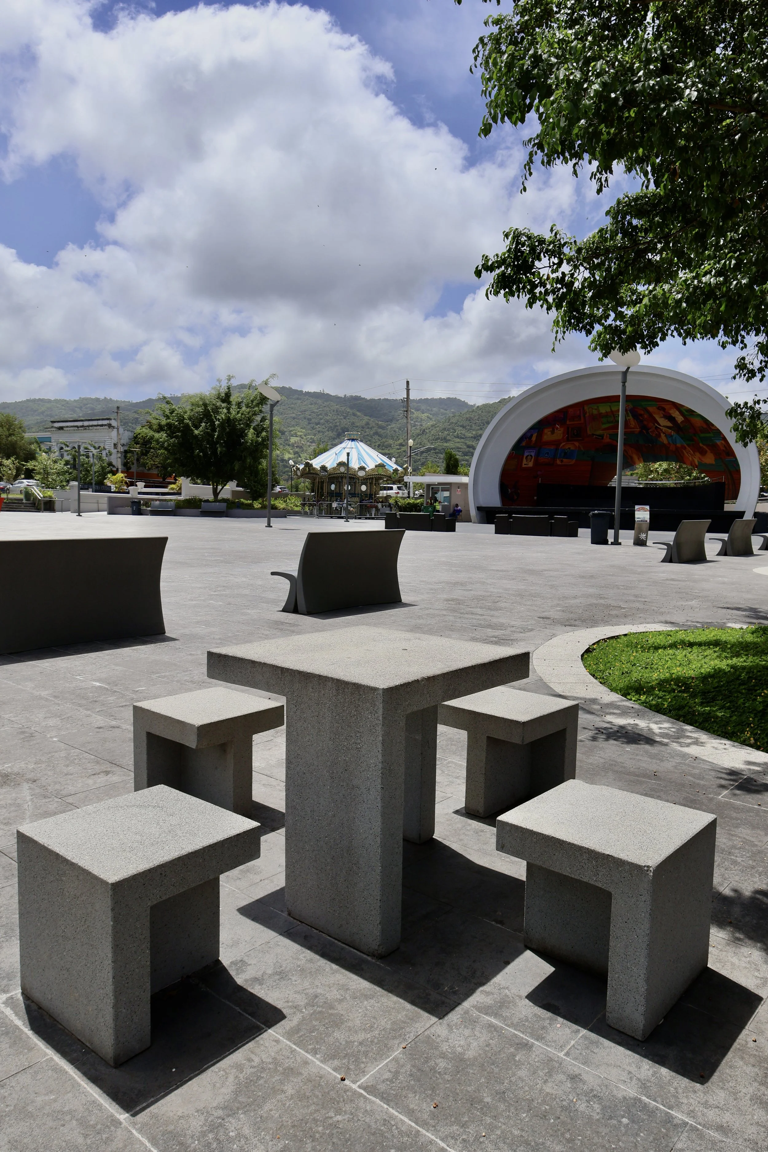 Outdoor plaza with concrete tables and benches, a stage with a curved white roof, trees, and a carousel in the background. Hills and cloudy sky are visible.