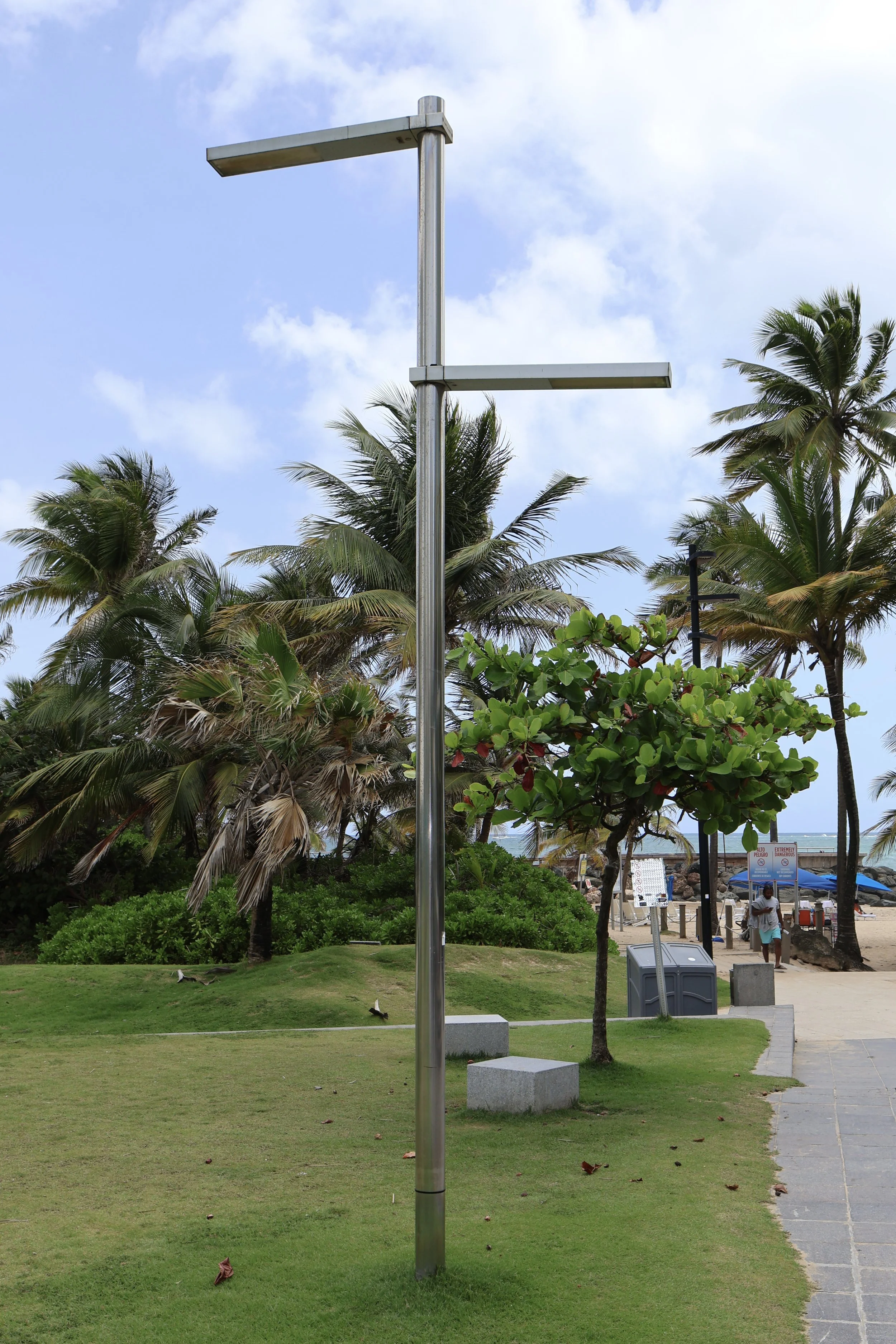 Empty outdoor sculpture structure with two horizontal beams on a tall metallic pole, situated on a grassy area near a beach with palm trees.