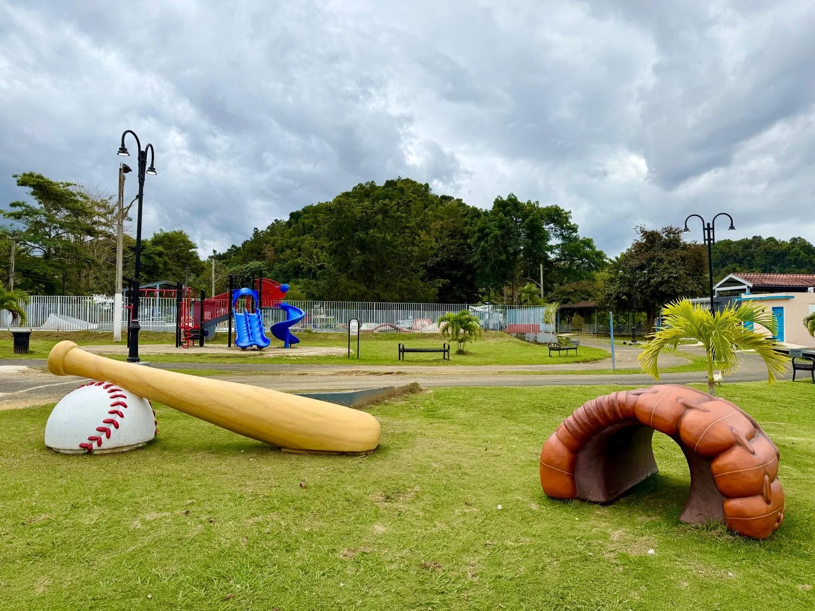 Children's playground with oversized baseball bat and baseball as decorative elements, benches, and a landscaping area with small palm trees, set against a backdrop of trees and cloudy sky.