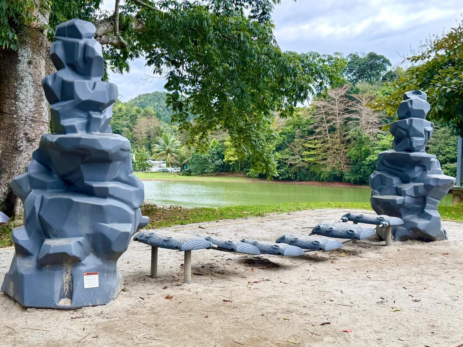 Outdoor park scene with two large, stylized gray rock sculptures and a wooden bench near a lake, surrounded by green trees and hills.